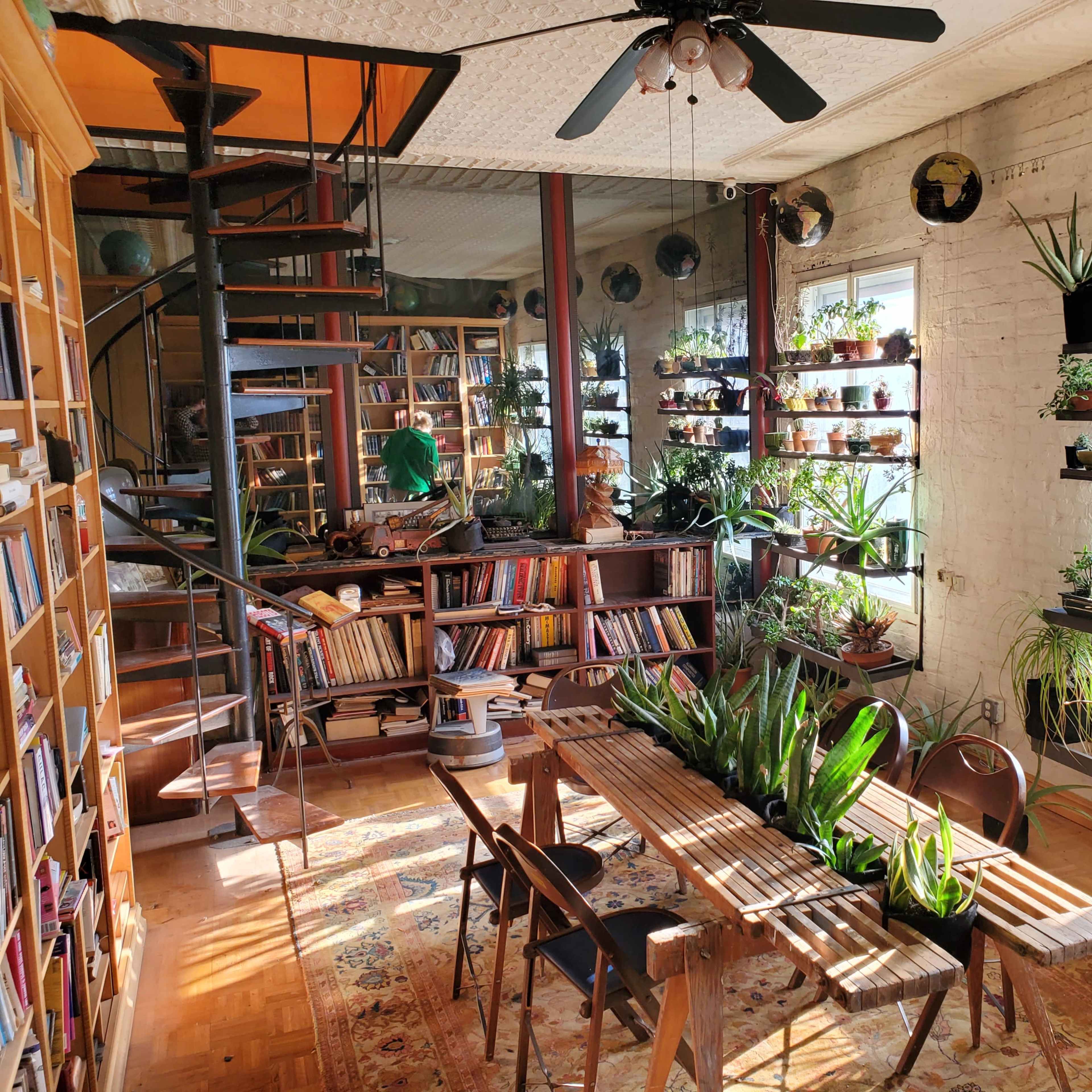 A cozy, plant-filled reading room with wooden shelves of books, a spiral staircase, and a wooden table surrounded by greenery.