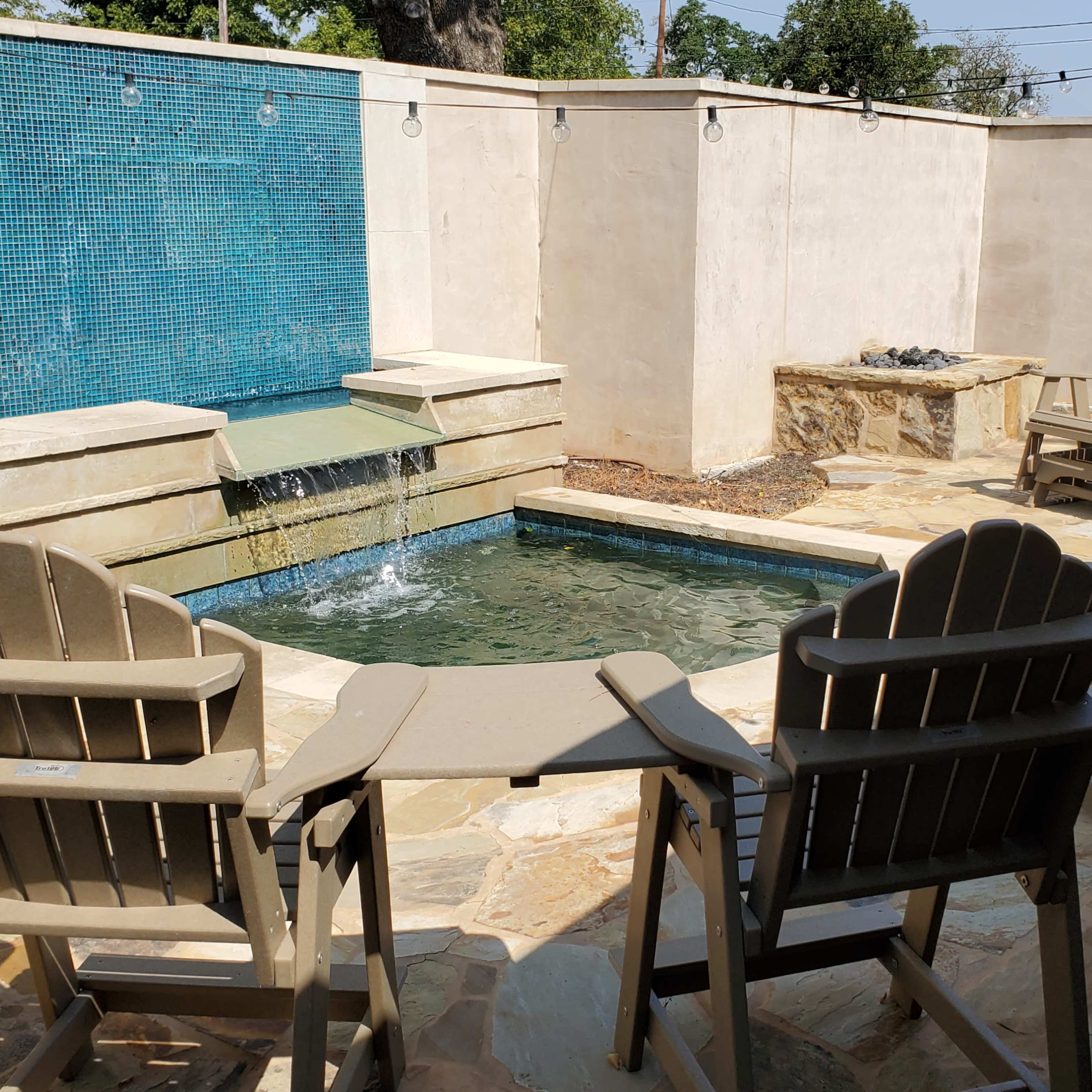 A small outdoor area featuring a shallow pool with a water fountain, surrounded by several Adirondack chairs on a stone patio.