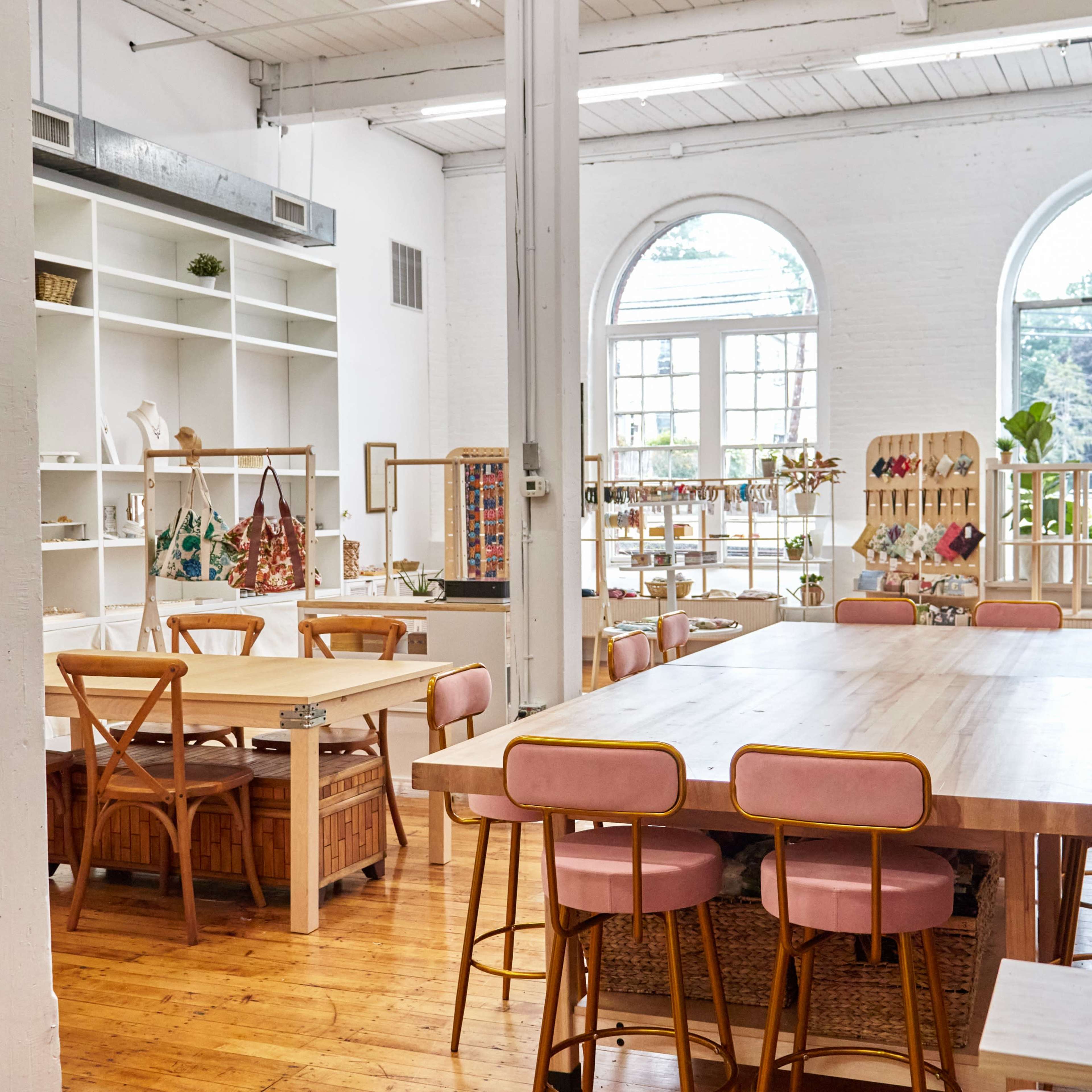 A spacious, bright room with large windows, featuring wooden tables and chairs arranged for a communal workspace.