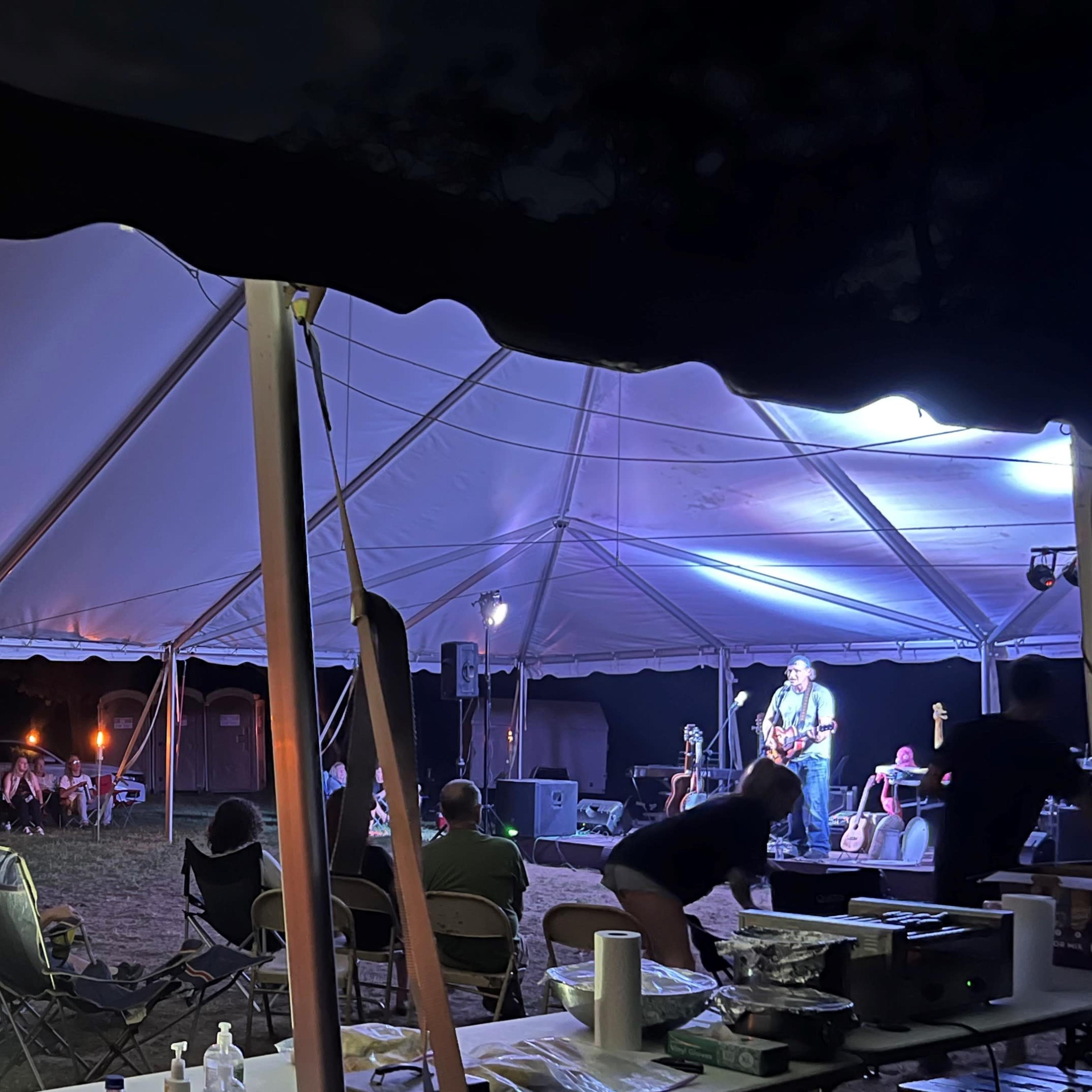 A live music performance is taking place under a large tent, with an audience seated in front of the stage.