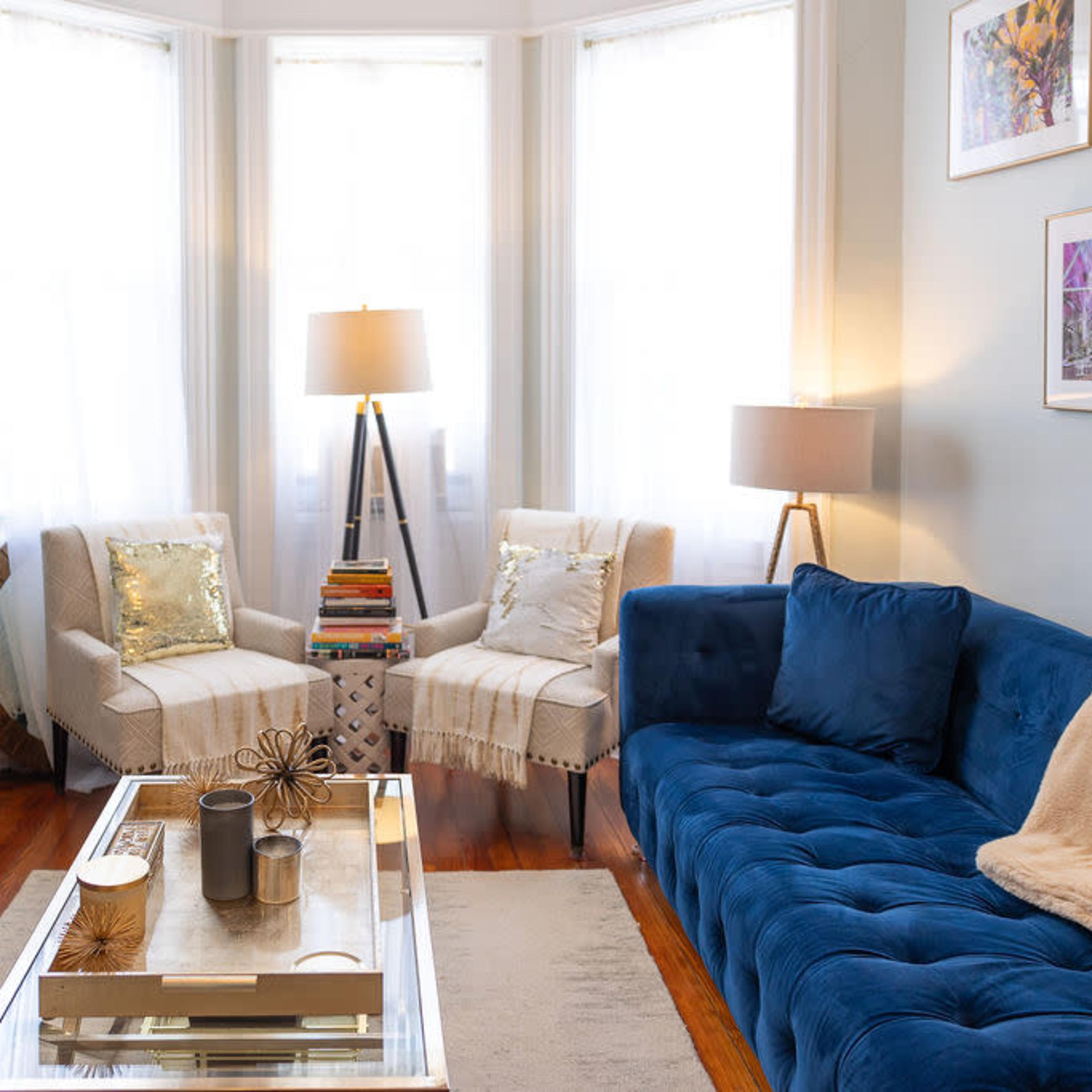 A cozy living room featuring a blue tufted sofa, two armchairs, a coffee table, and large windows allowing natural light to fill the space.