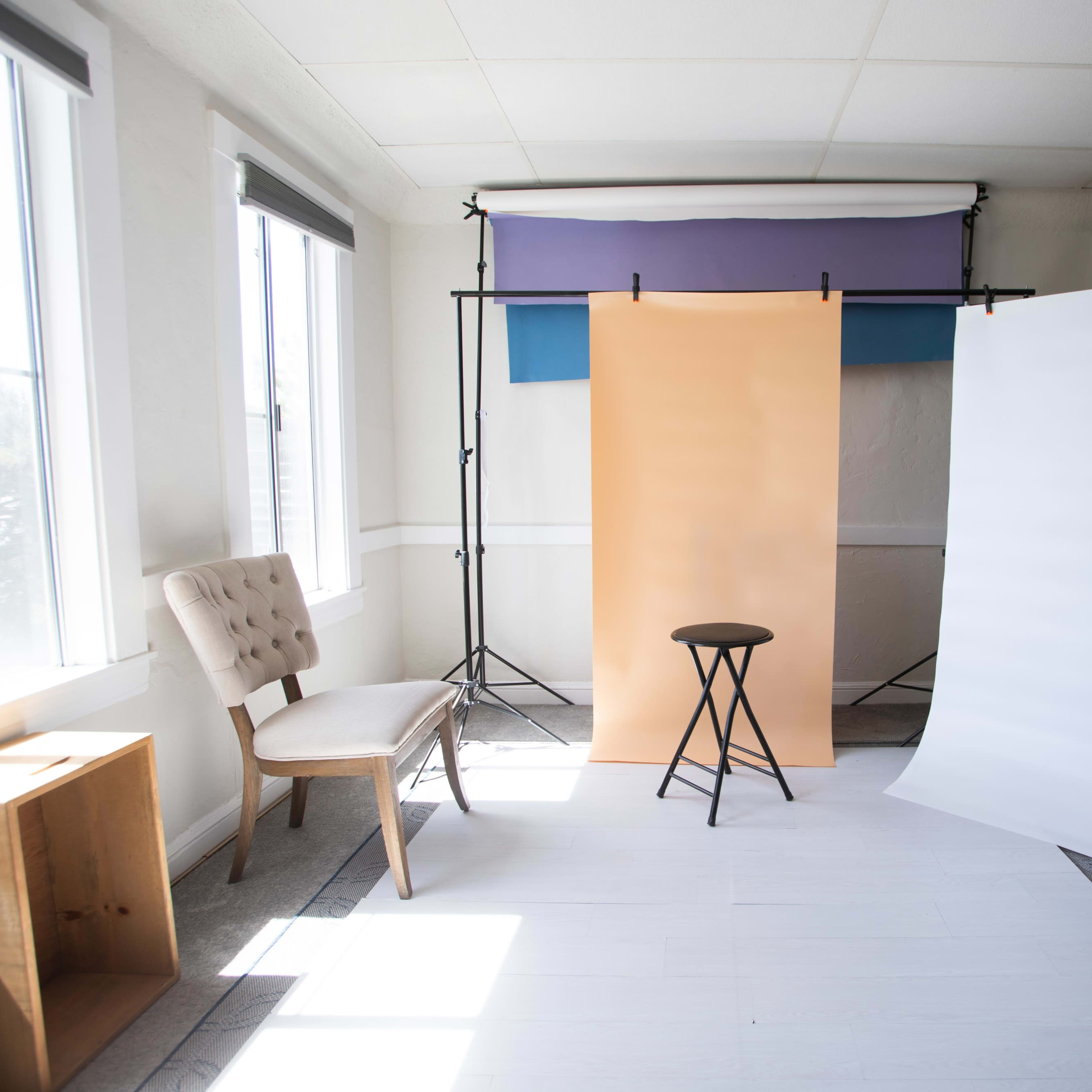 A photography studio setup with a stool in front of three backdrops: one orange, one white, and one purple, along with a chair and natural light coming through the windows.