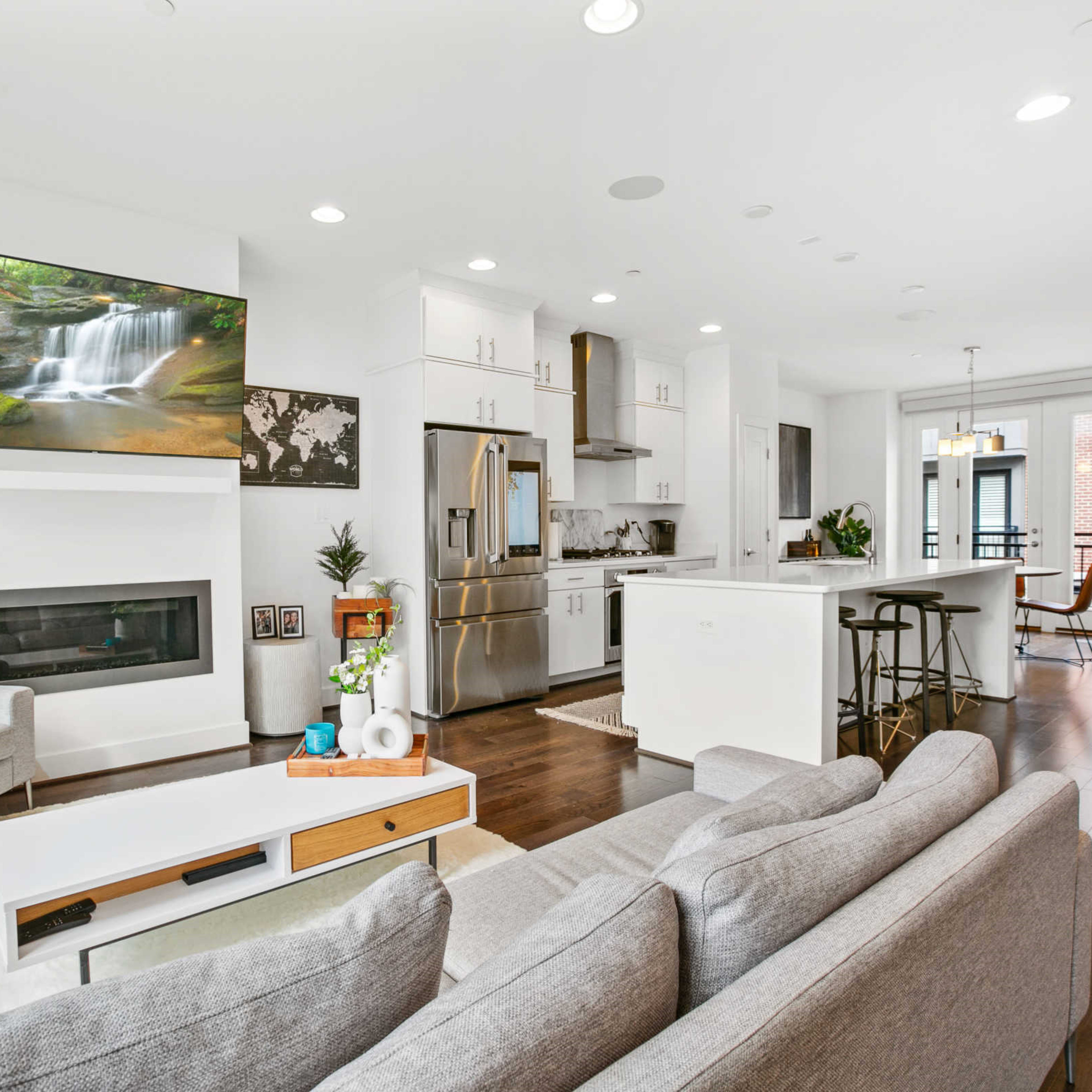 A modern living room and kitchen area featuring a large television mounted on the wall, a sofa, a dining table, and a balcony view.