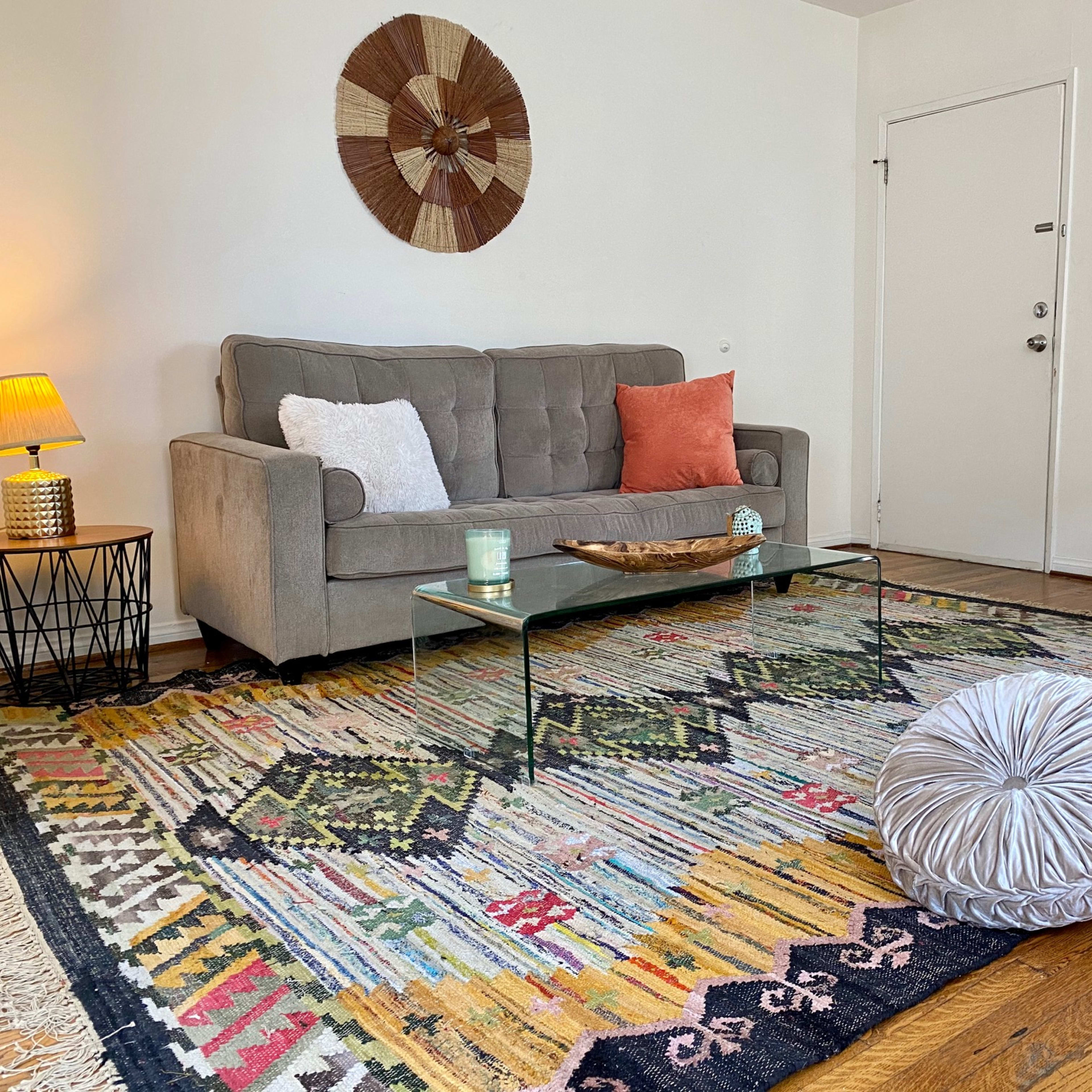 The scene shows a cozy living room with a grey sofa, a glass coffee table, a patterned area rug, a decorative wall hanging, and an orange accent chair.