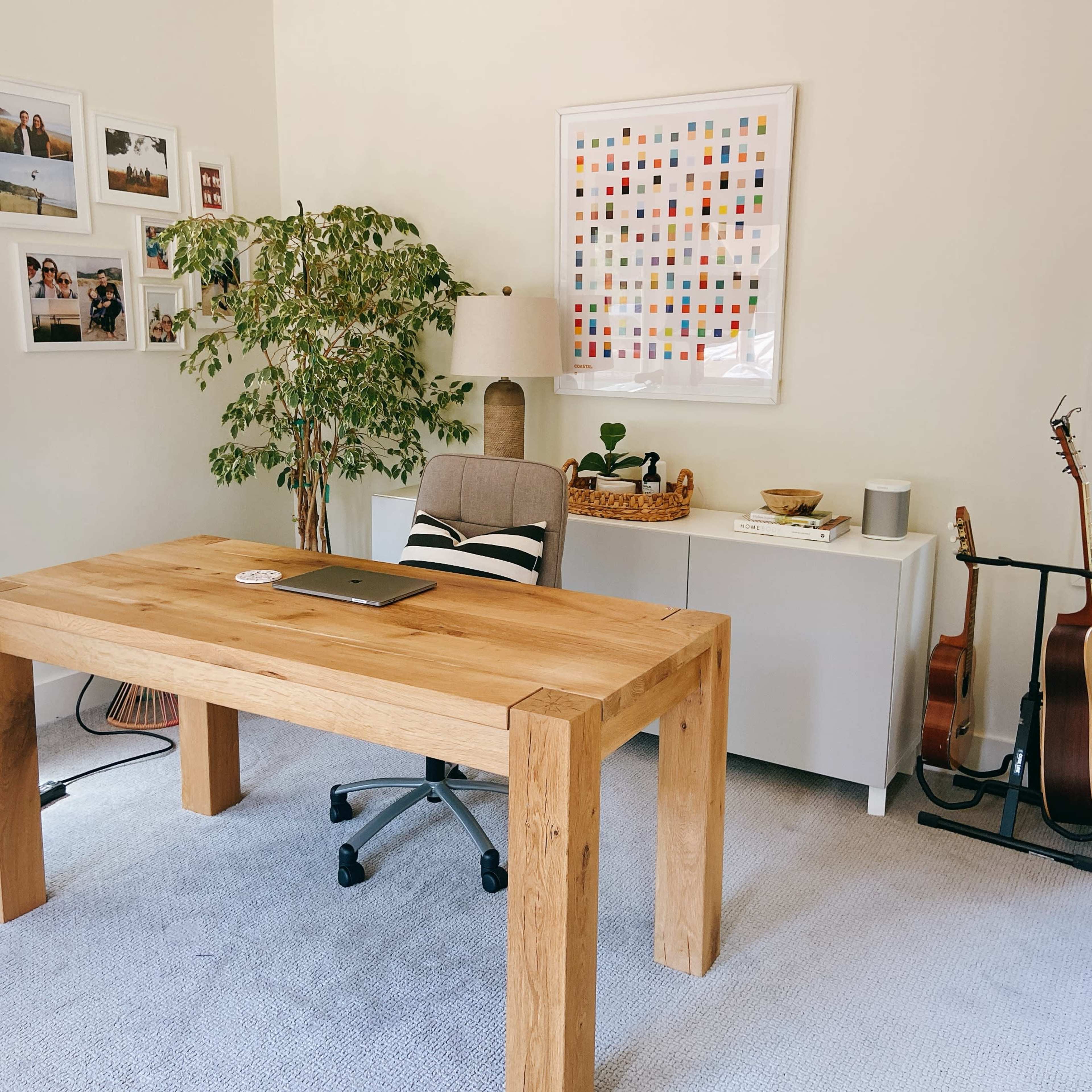 A wooden desk with a laptop sits in a brightly lit office featuring a potted plant, a framed art piece on the wall, and a guitar support stand.