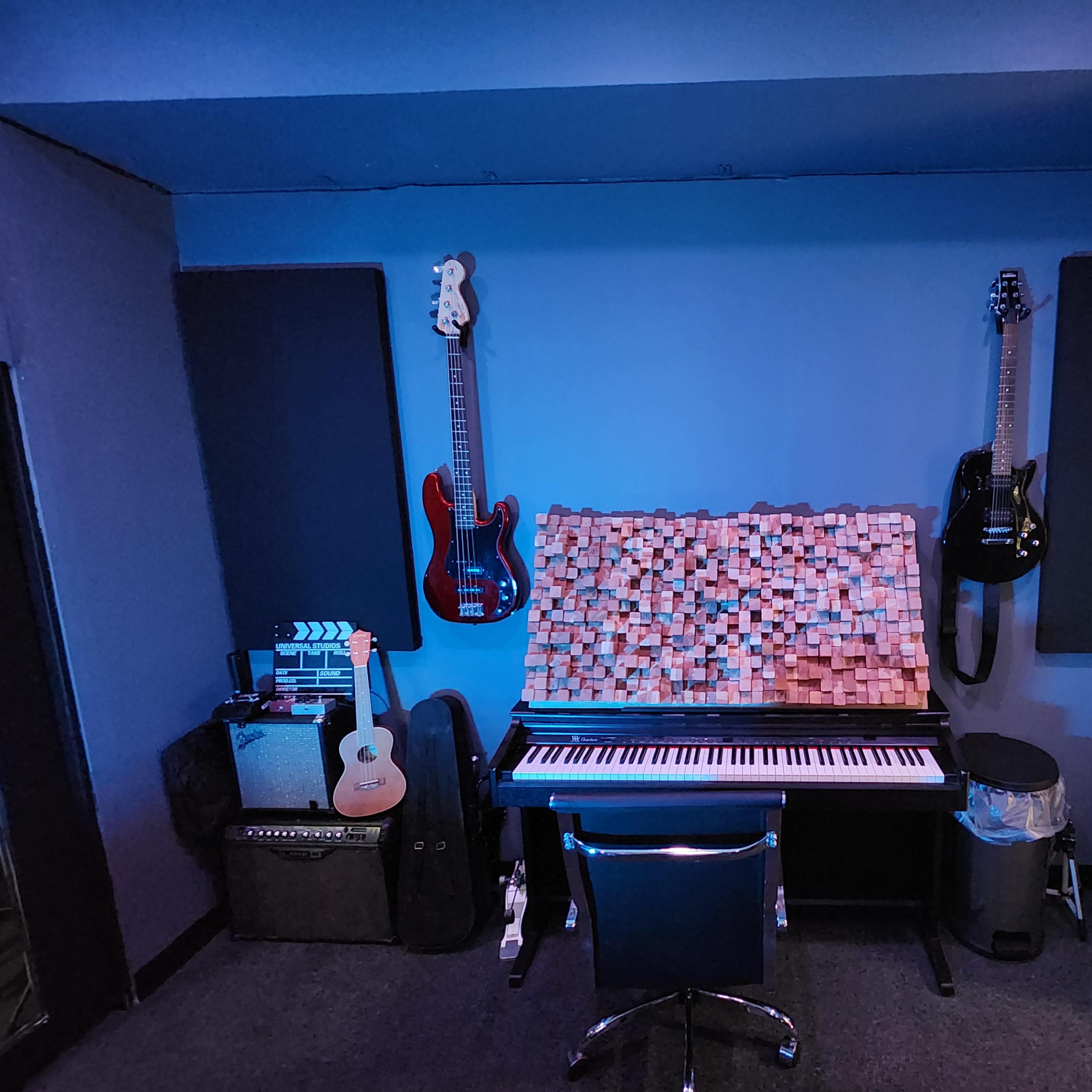 A music studio setup featuring a keyboard at the center, surrounded by various guitars hung on the walls, acoustic panels, and amplifiers.