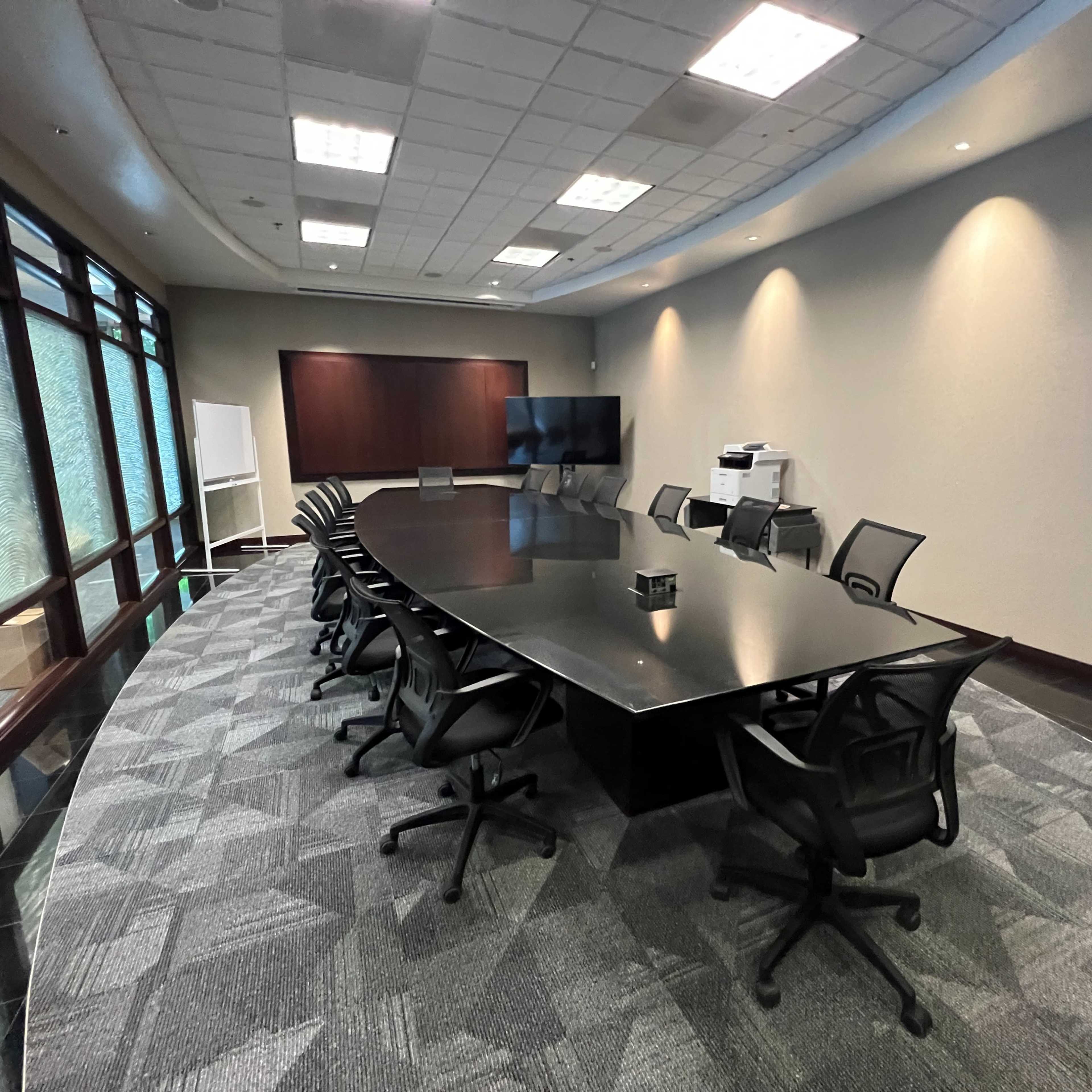 A modern conference room features a large black table surrounded by mesh office chairs, with a television screen and whiteboard against one wall.