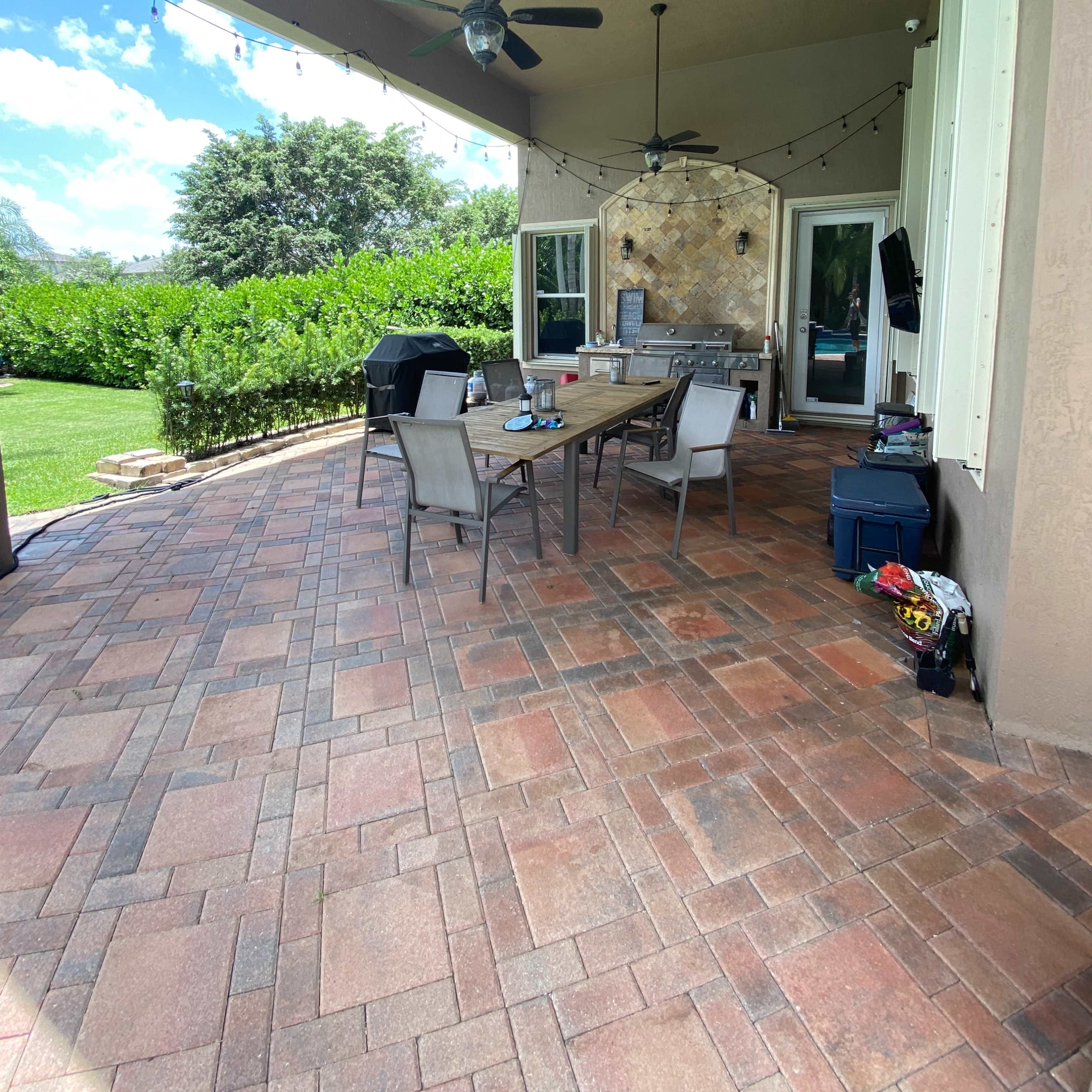 A covered patio features a long table surrounded by chairs, with a grill and outdoor seating area in a landscaped garden.