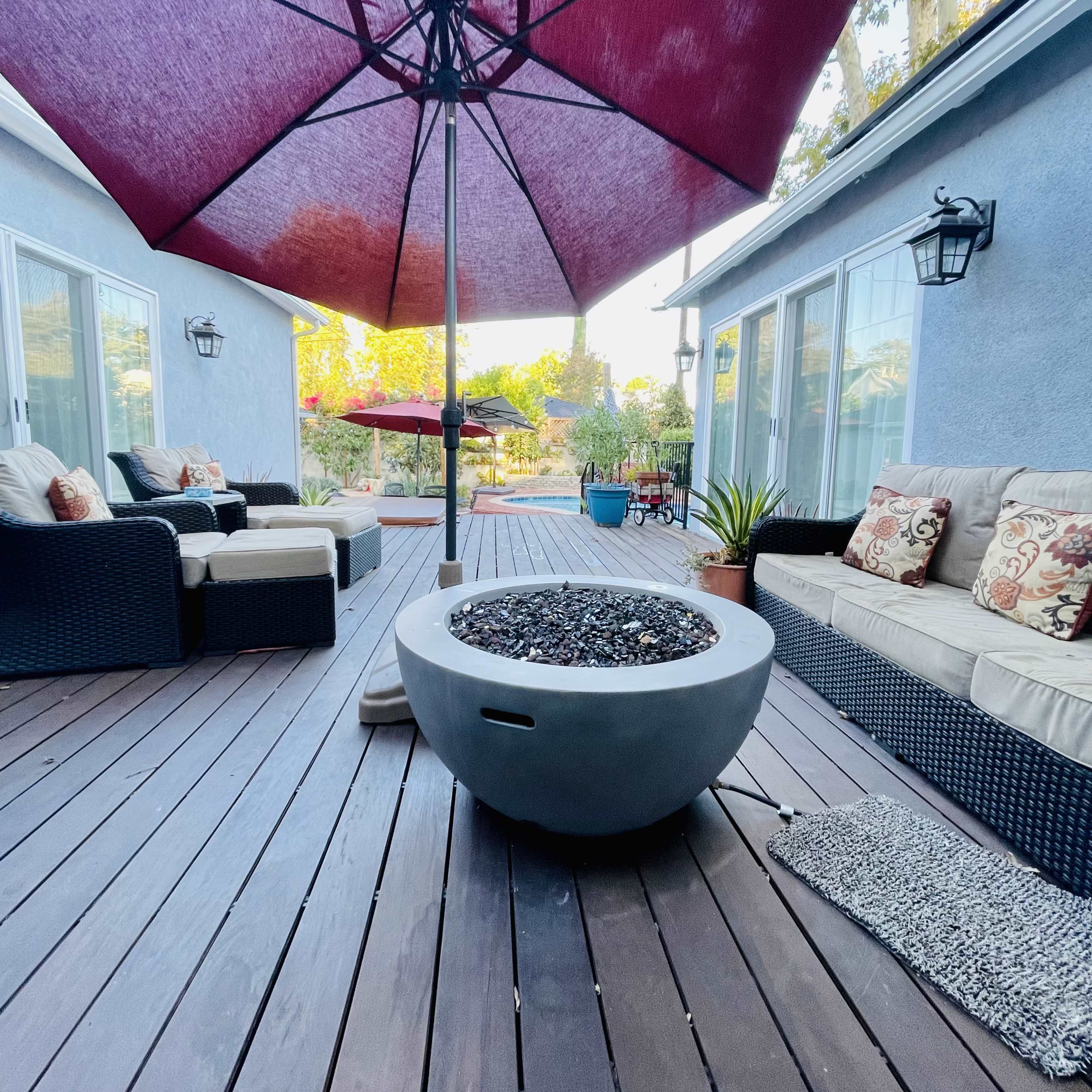 A patio area features a large umbrella over a round fire pit surrounded by outdoor seating and plants.