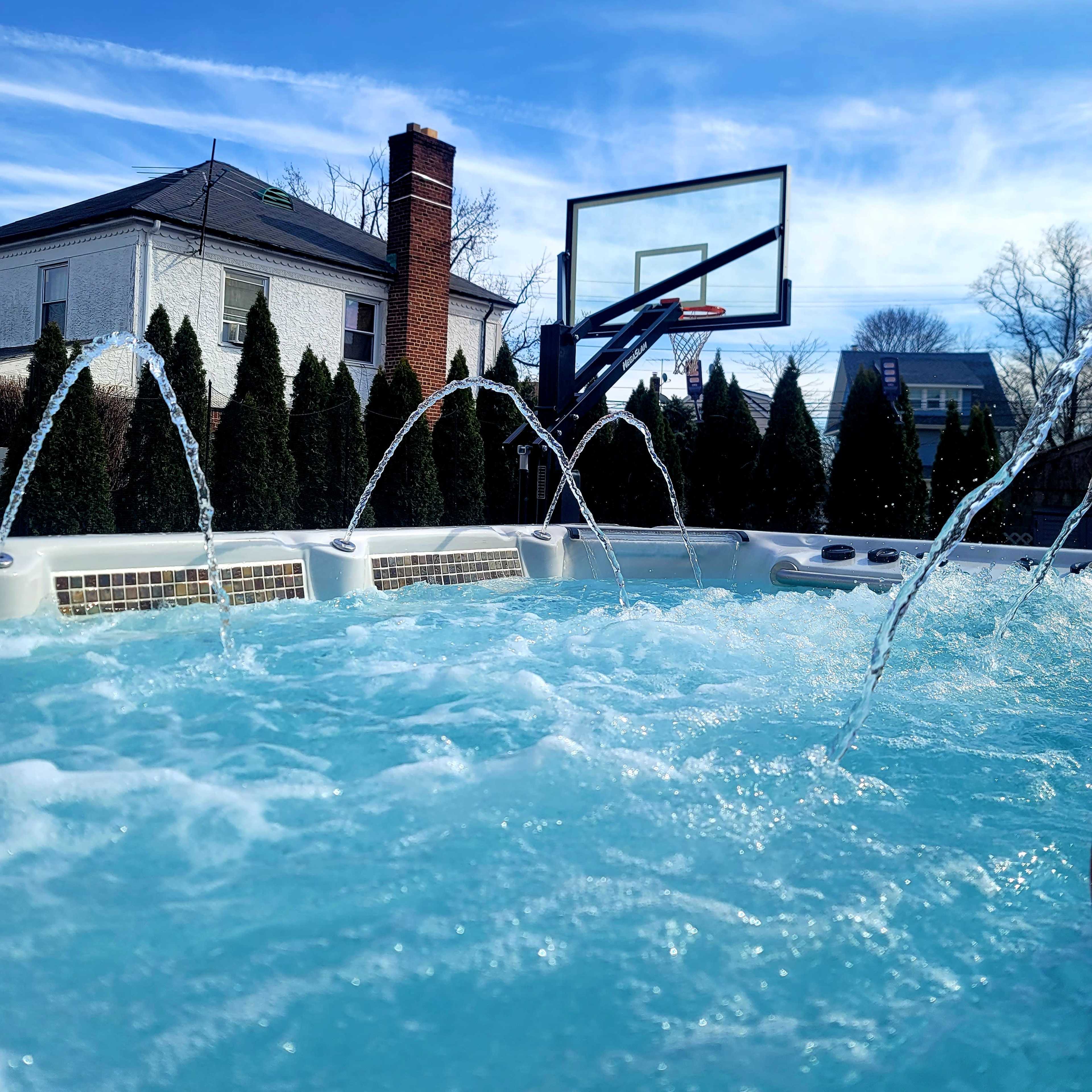 A hot tub with water jets and a basketball hoop in the background is set against a clear sky and a house surrounded by tall hedges.