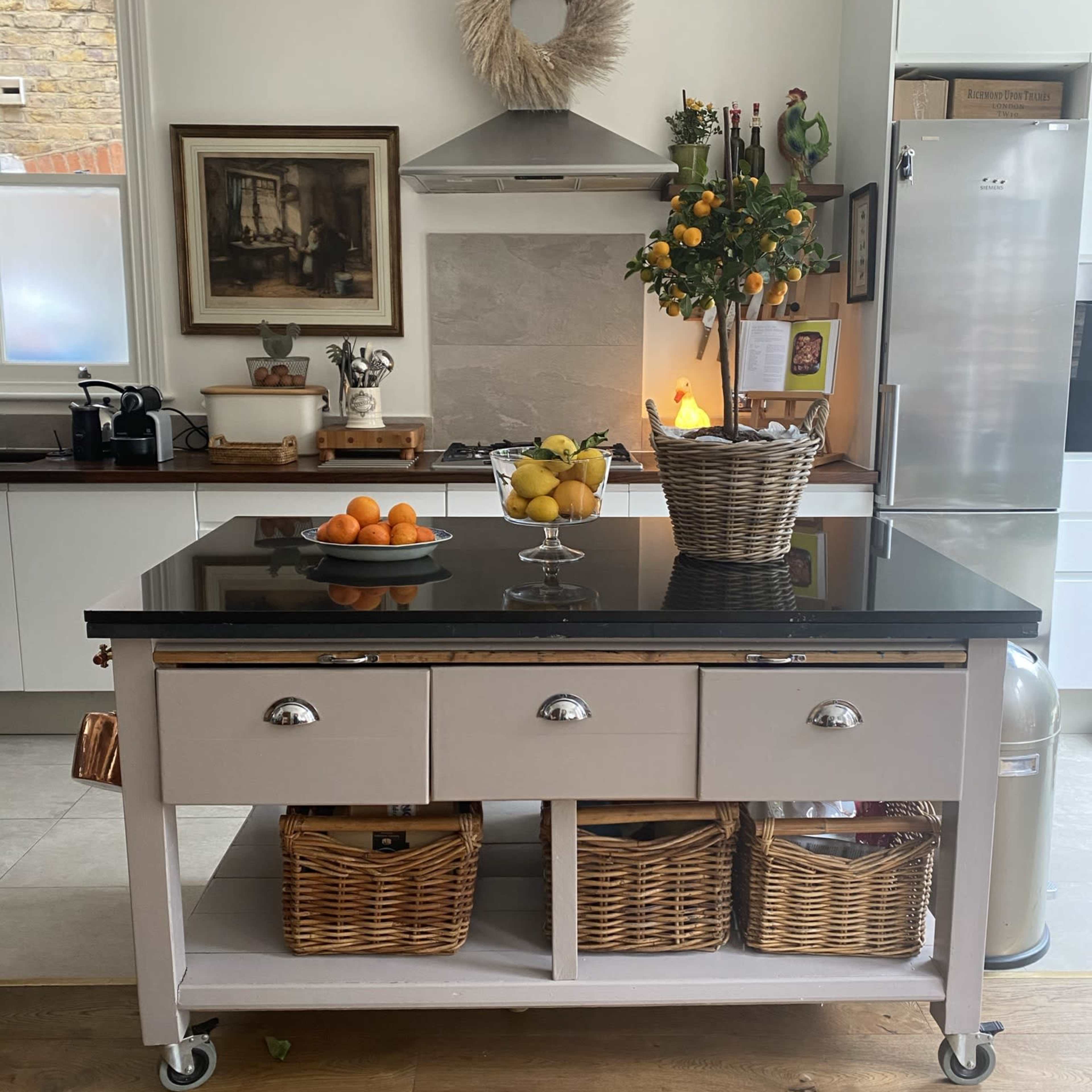 A modern kitchen features a central island with a fruit display, stainless steel appliances, and a view into a dining area with a wooden table.