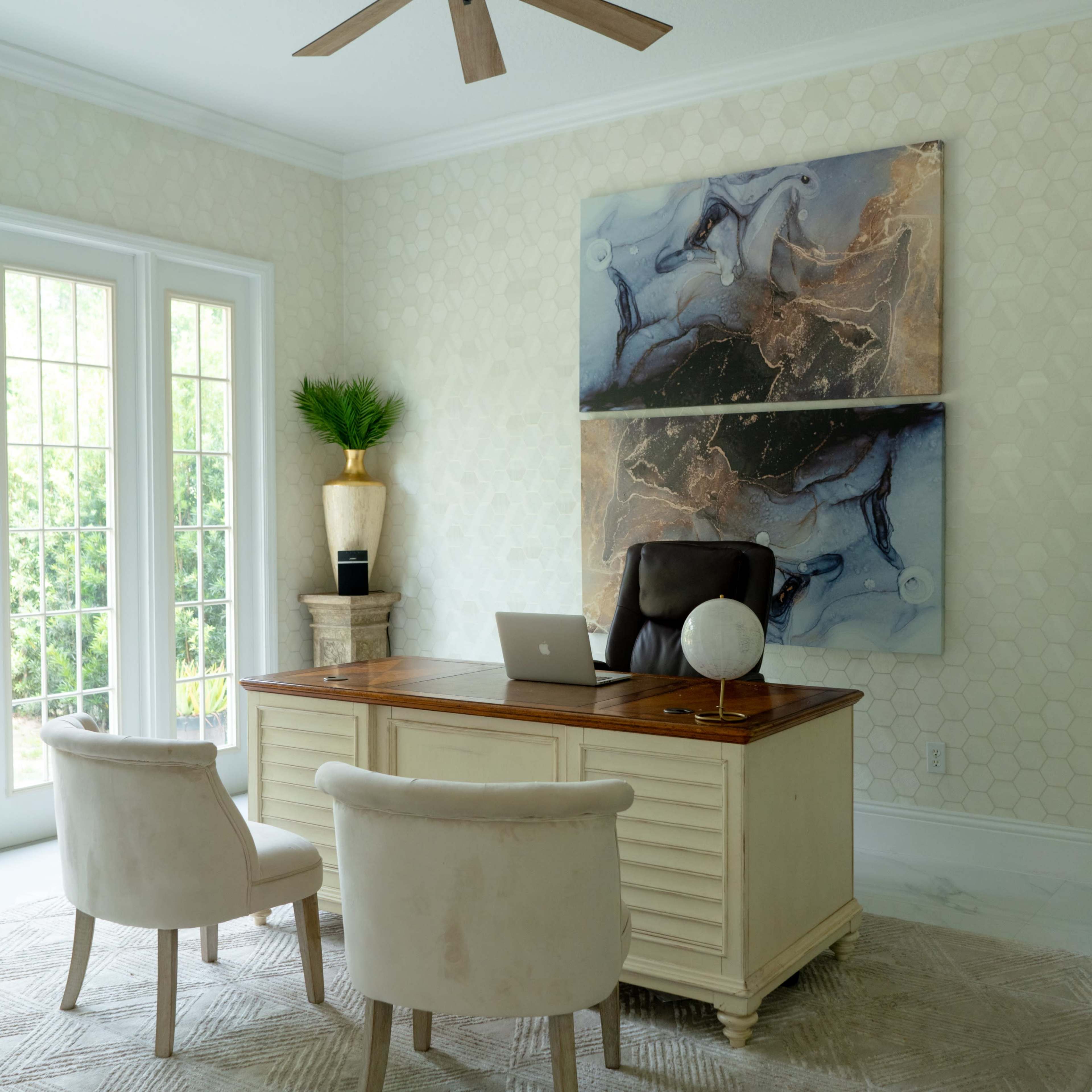 A well-decorated office features a wooden desk with a laptop, two upholstered chairs, and large windows allowing natural light to enter.