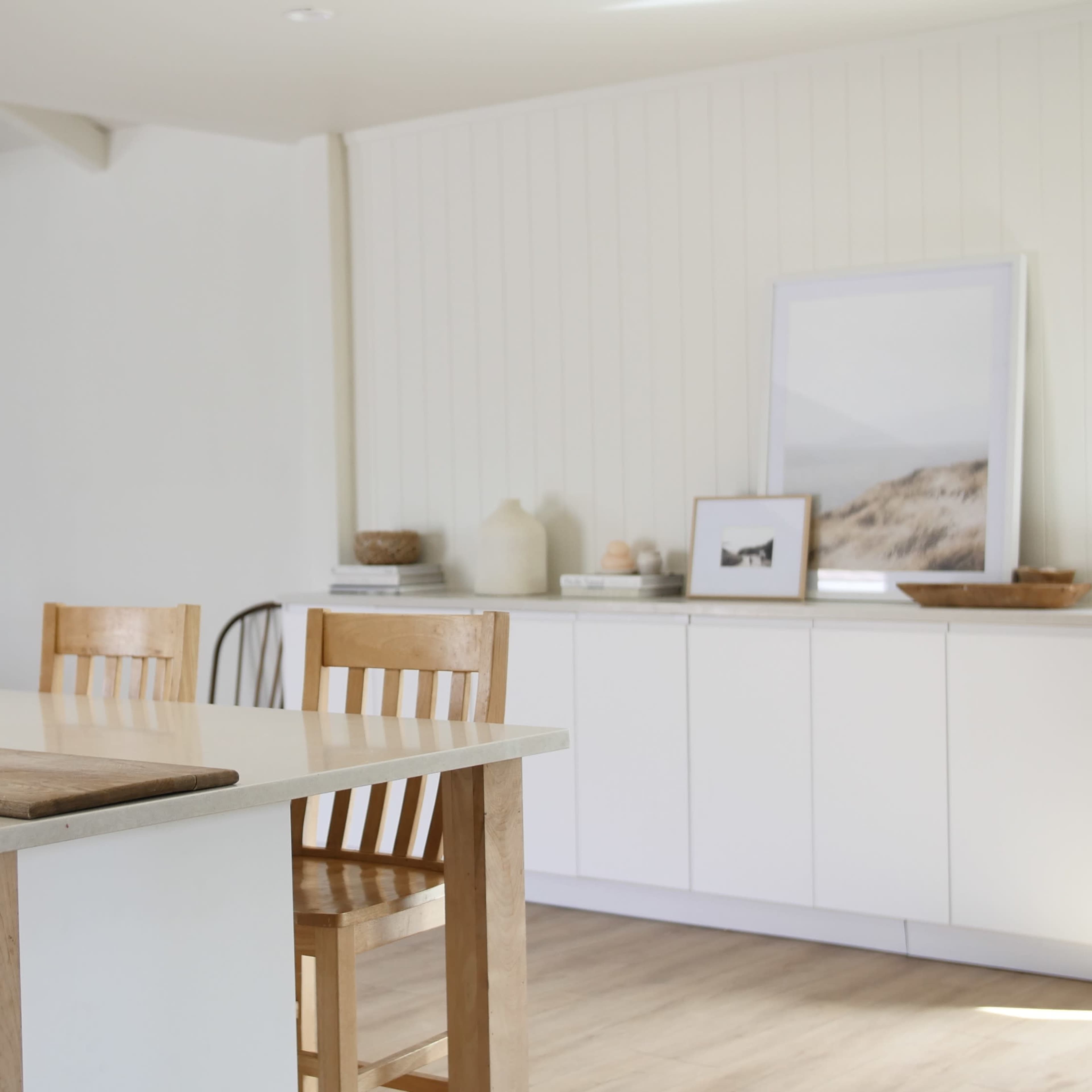 A minimalist kitchen with a wooden dining table and chairs, a stone countertop, and white cabinetry against a light-colored wall.