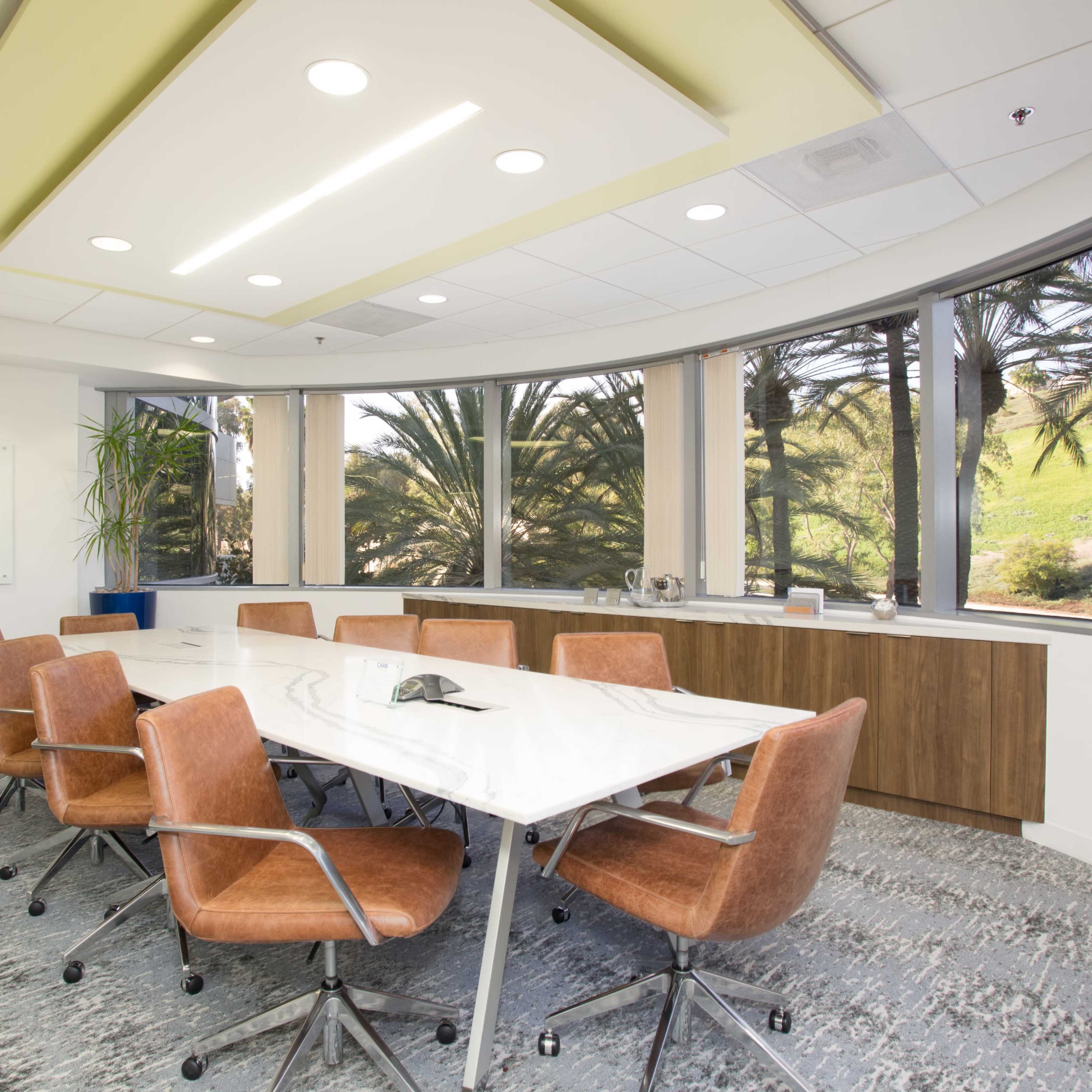 A conference room features a long white table surrounded by brown chairs, with large windows overlooking palm trees.