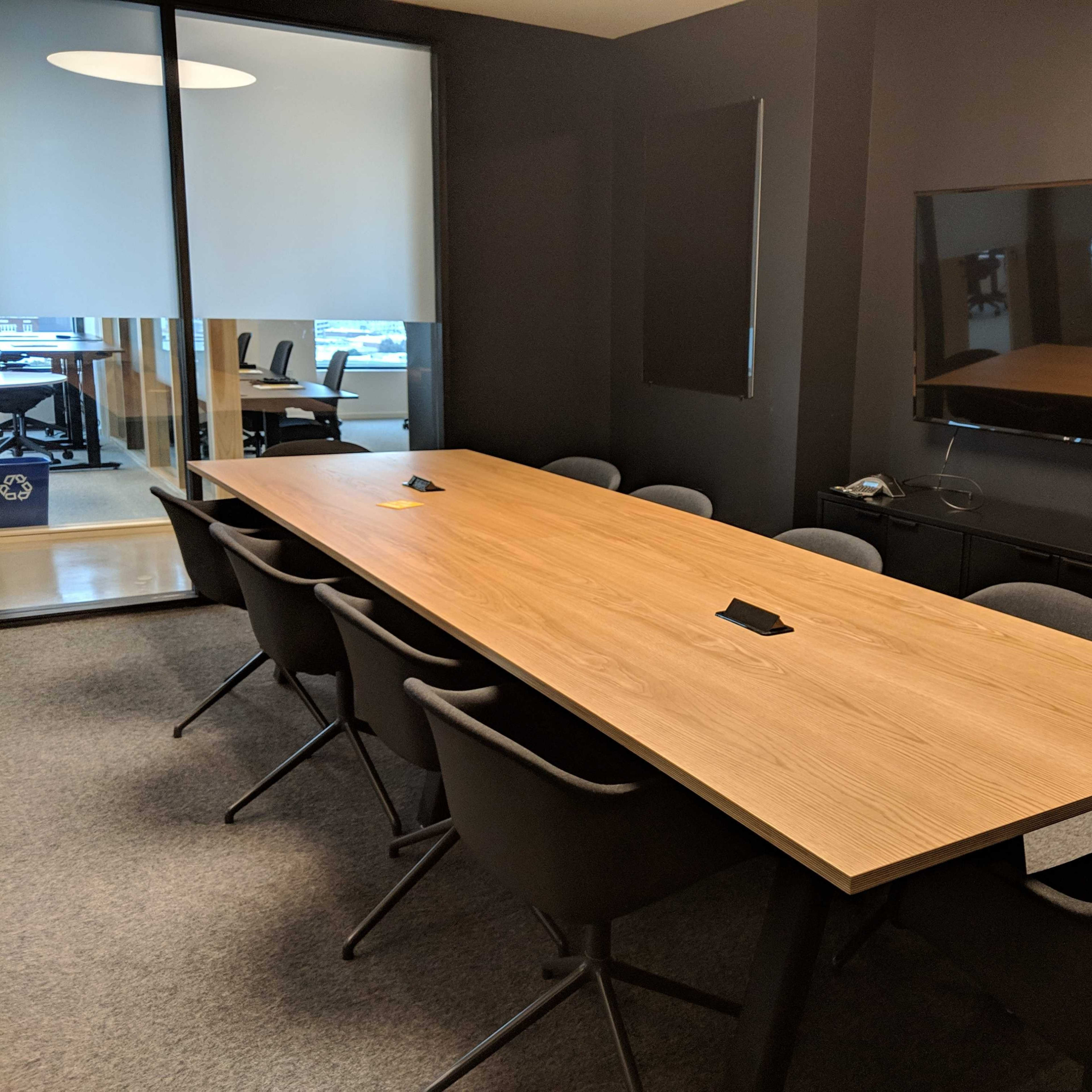 A large wooden conference table with black chairs is positioned in a dark conference room, adjacent to a glass-walled area with additional meeting space.