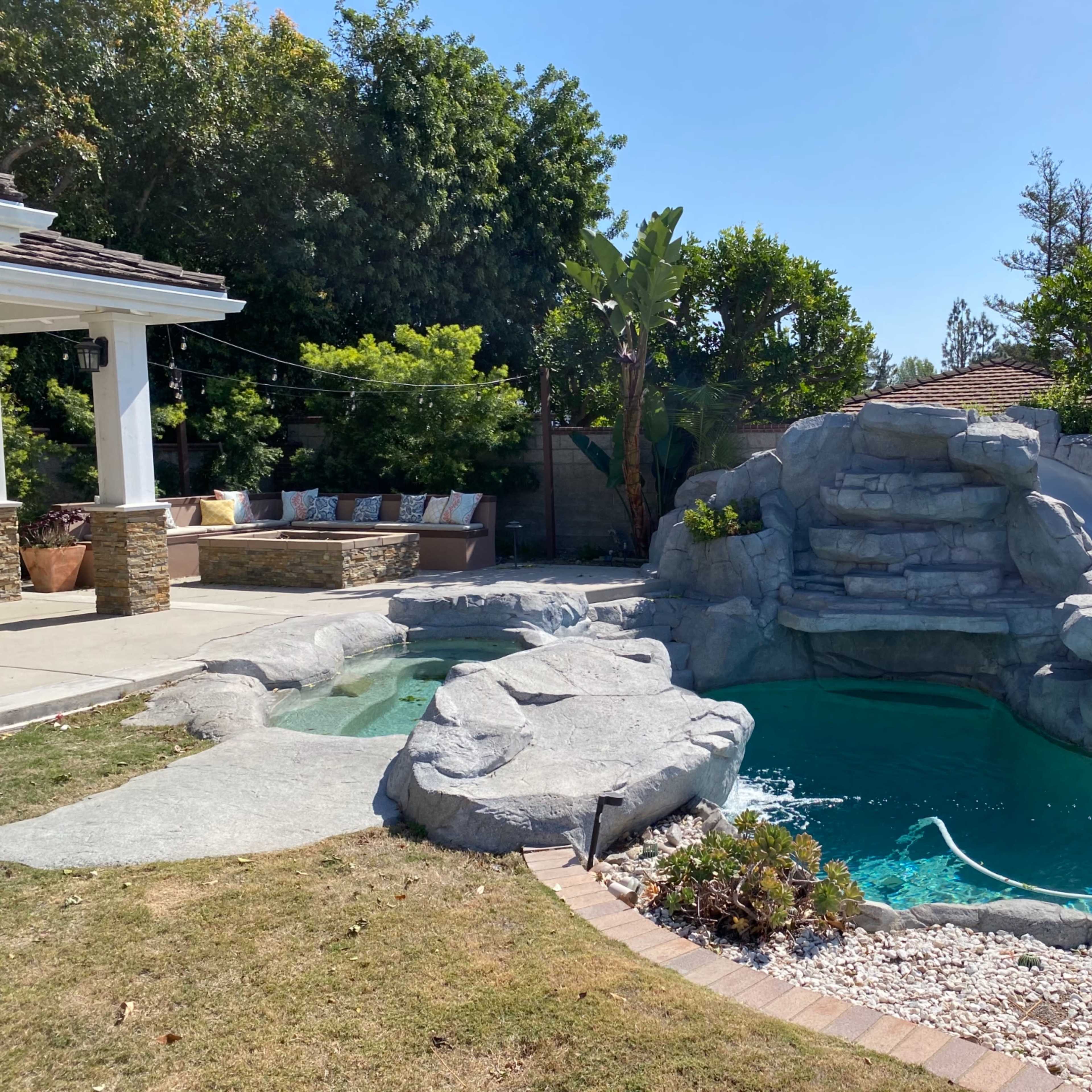 A backyard with a swimming pool featuring a rock formation and a water slide, surrounded by greenery and patio seating.