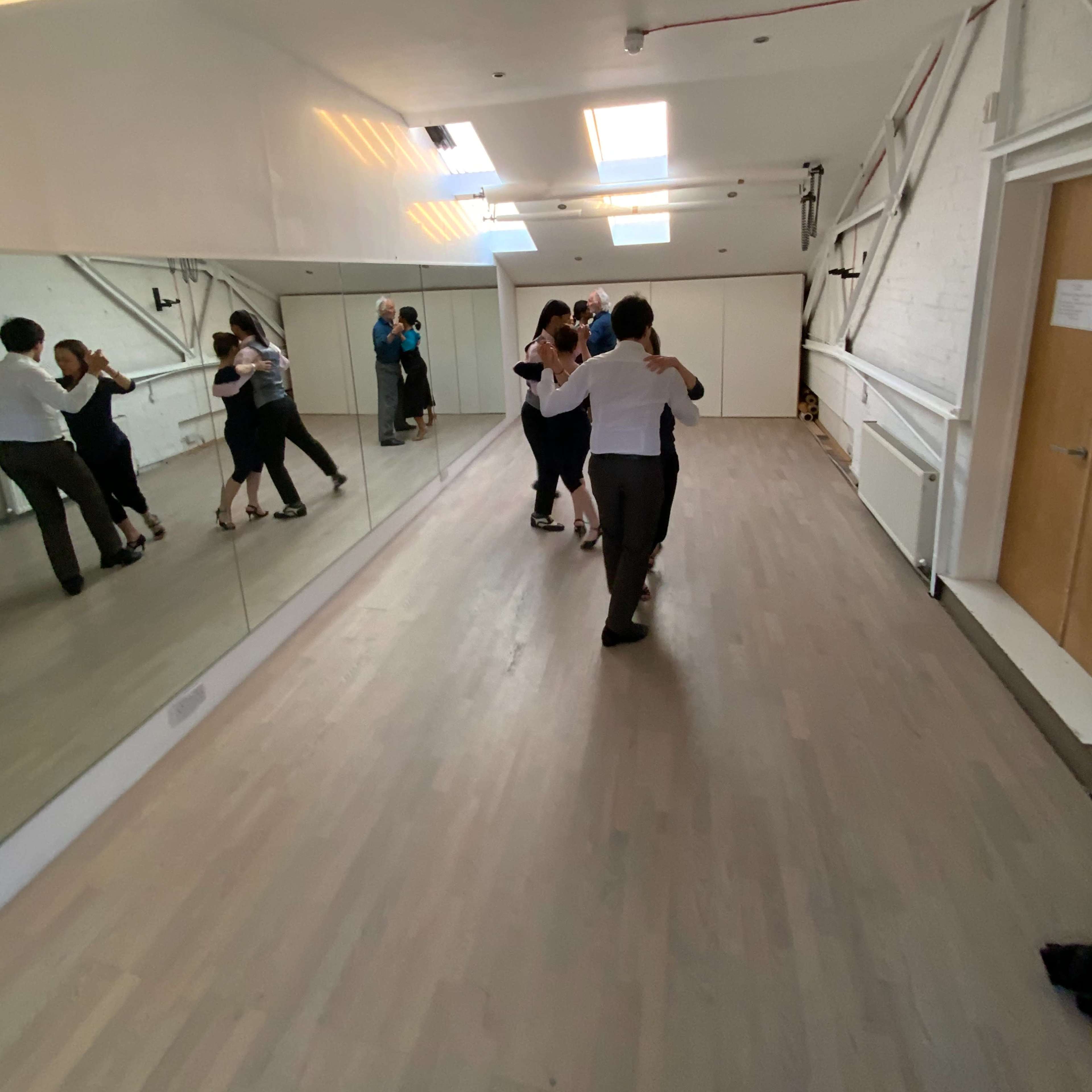 A dance studio features couples practicing in front of a large mirror, with wooden flooring and skylights above.