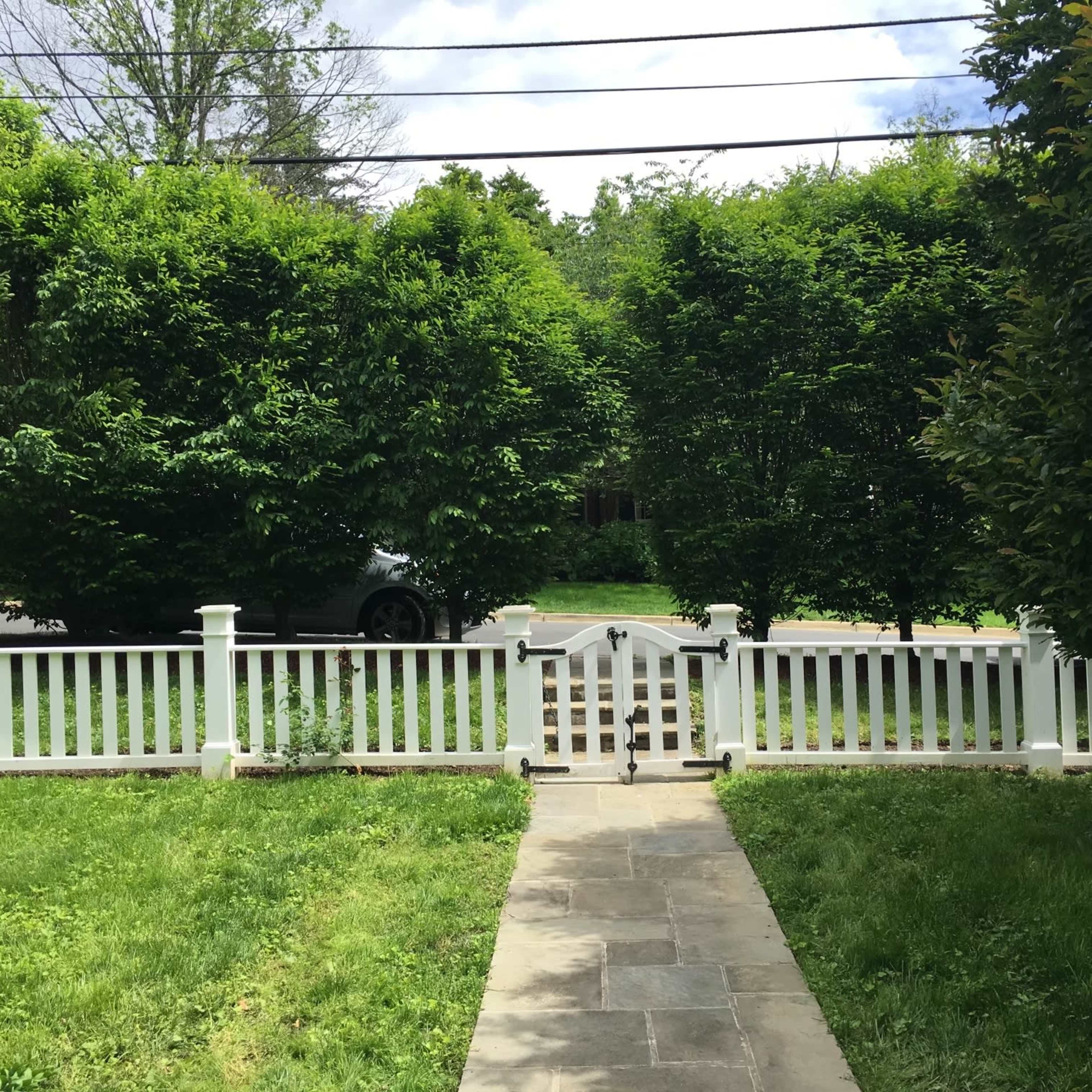 A walkway leads through a white picket fence gate to a grassy yard surrounded by tall hedges and trees.