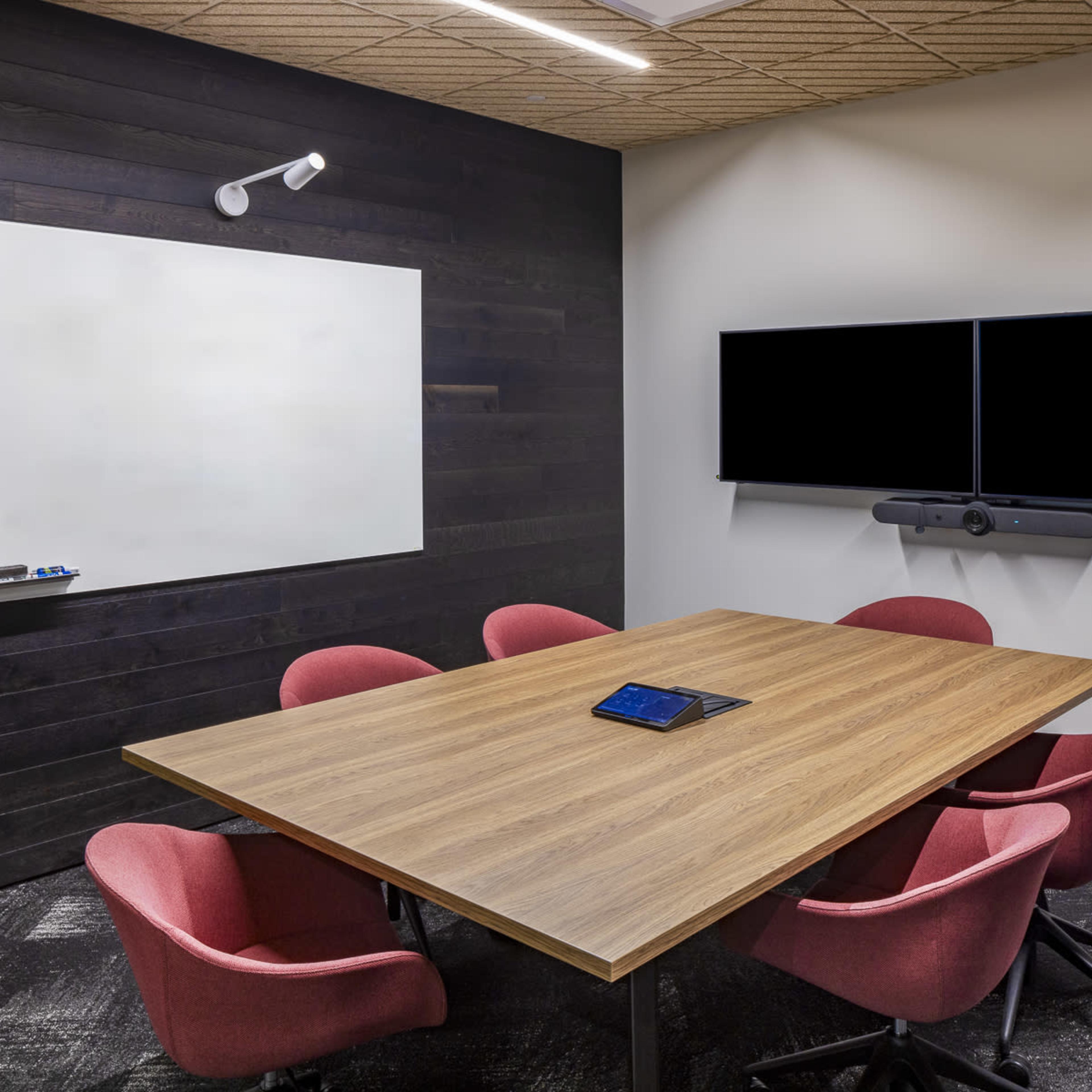 A modern conference room features a wooden table surrounded by red chairs, a whiteboard on the wall, and two large screens mounted on the opposite wall.