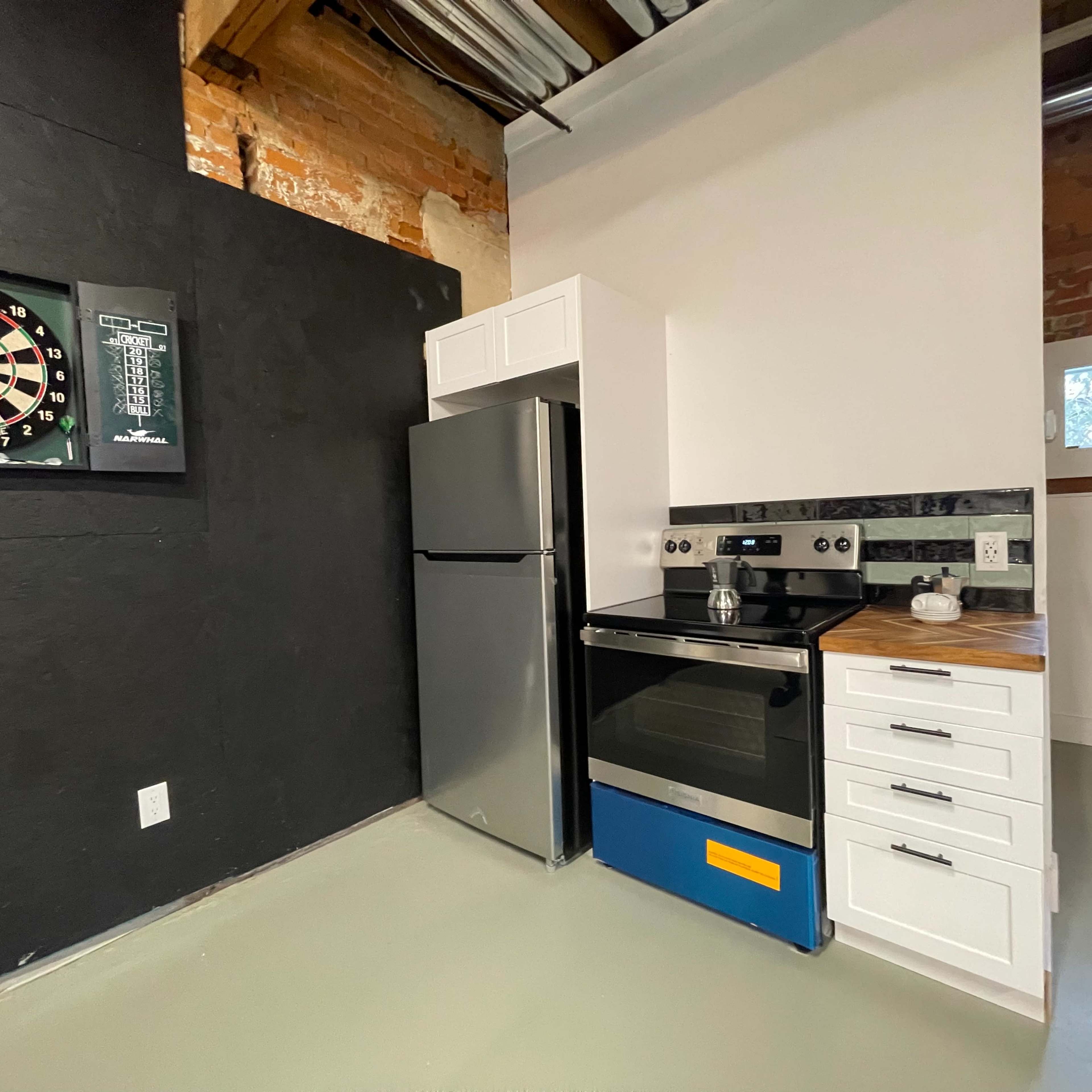 A modern kitchen corner featuring a stainless steel refrigerator, an oven with a blue accent, and a dartboard mounted on a black wall.