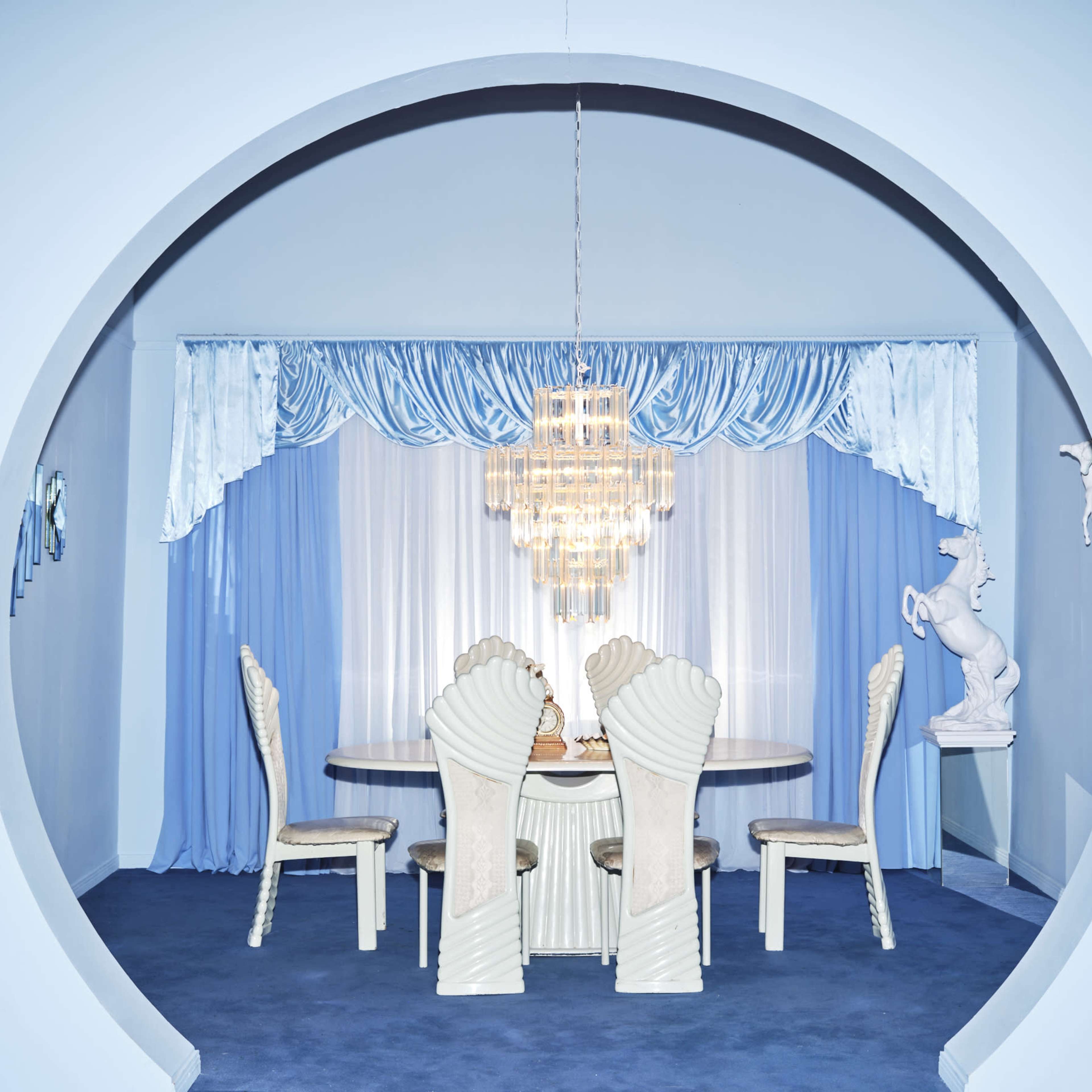 A circular arch frames a dining area with a round table and chairs, illuminated by a chandelier, surrounded by light blue curtains.