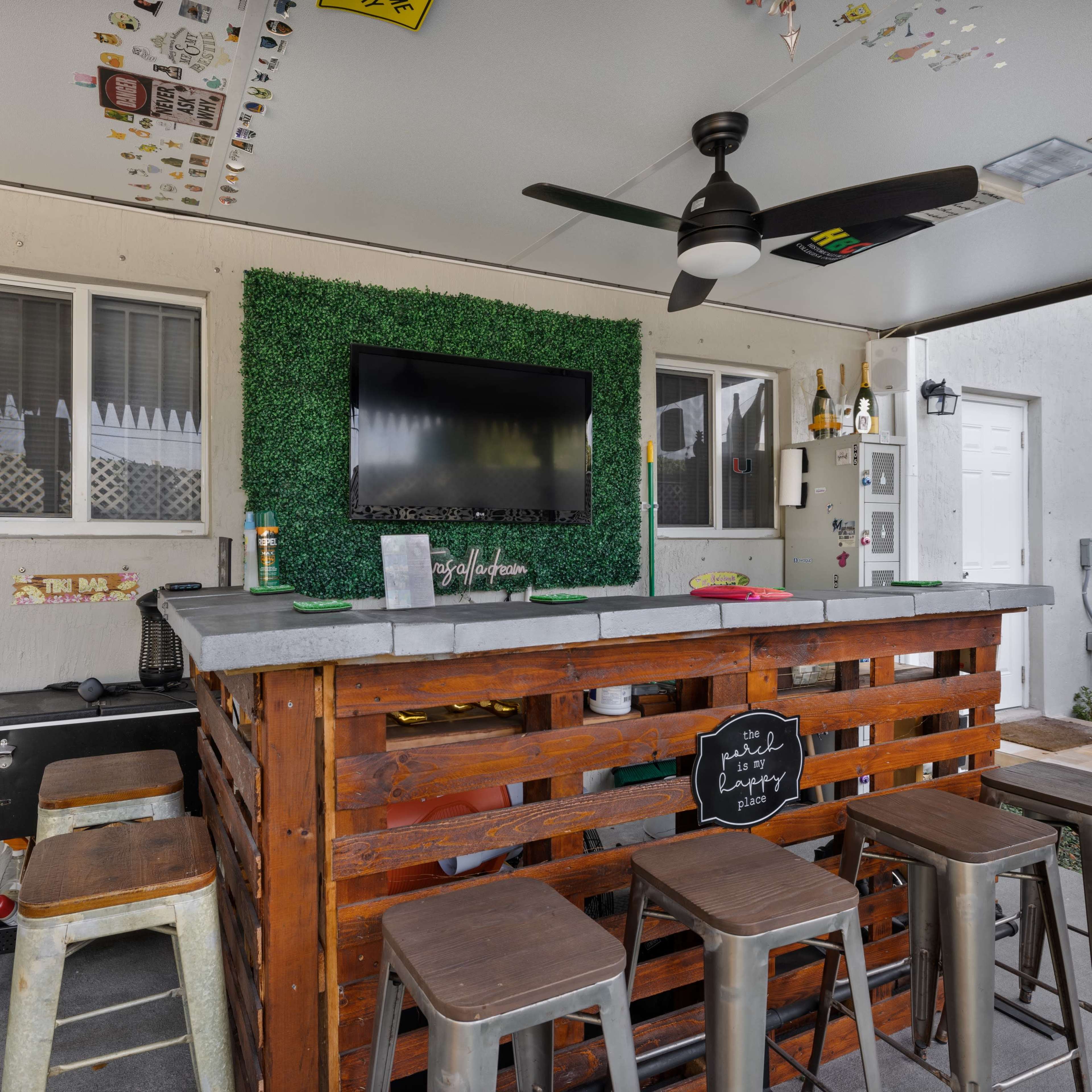 An outdoor bar area with a wooden counter, metal stools, a television mounted on the wall, and a dartboard on the side.