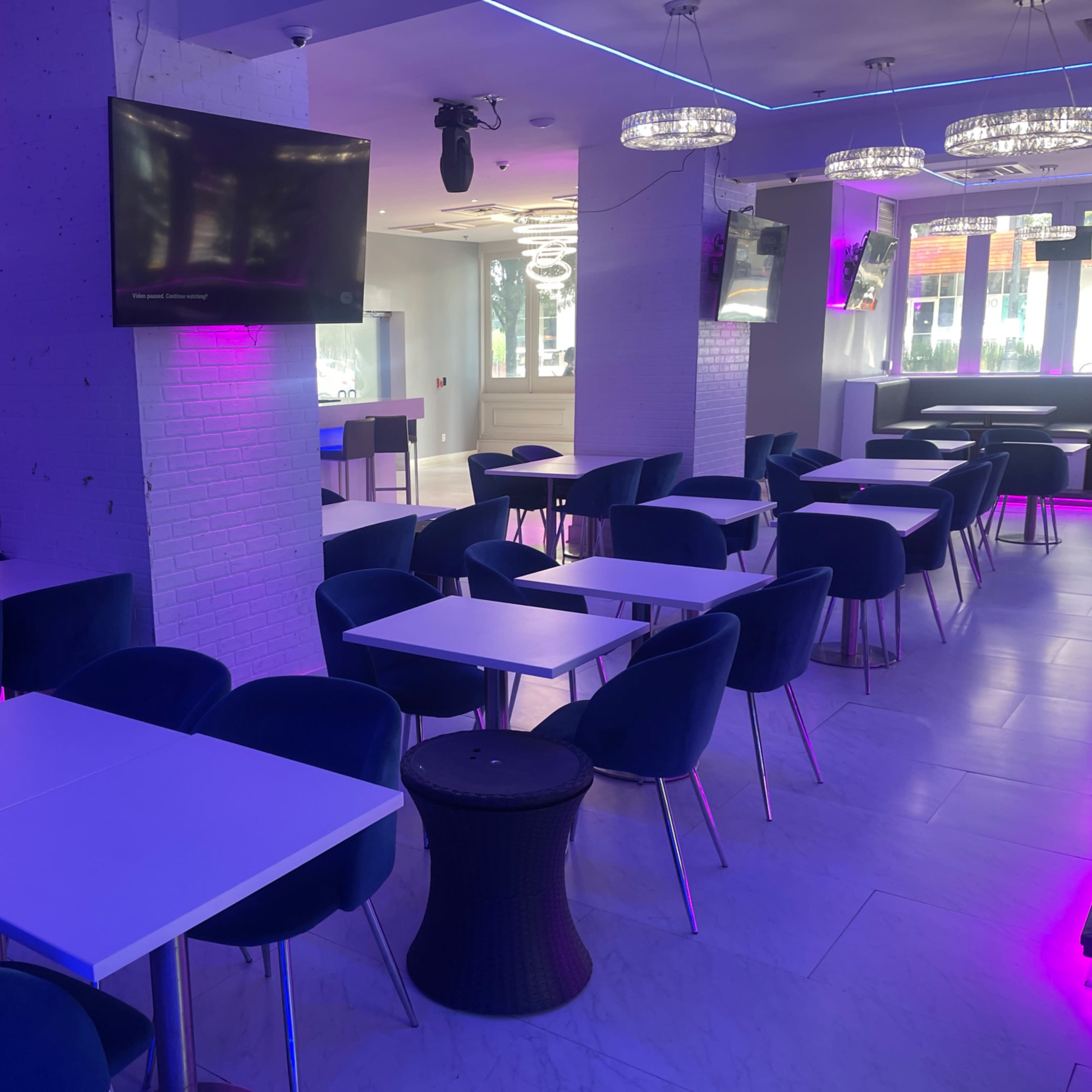 An empty dining area with blue chairs and white tables, illuminated by purple and white lighting, and a bar in the background.