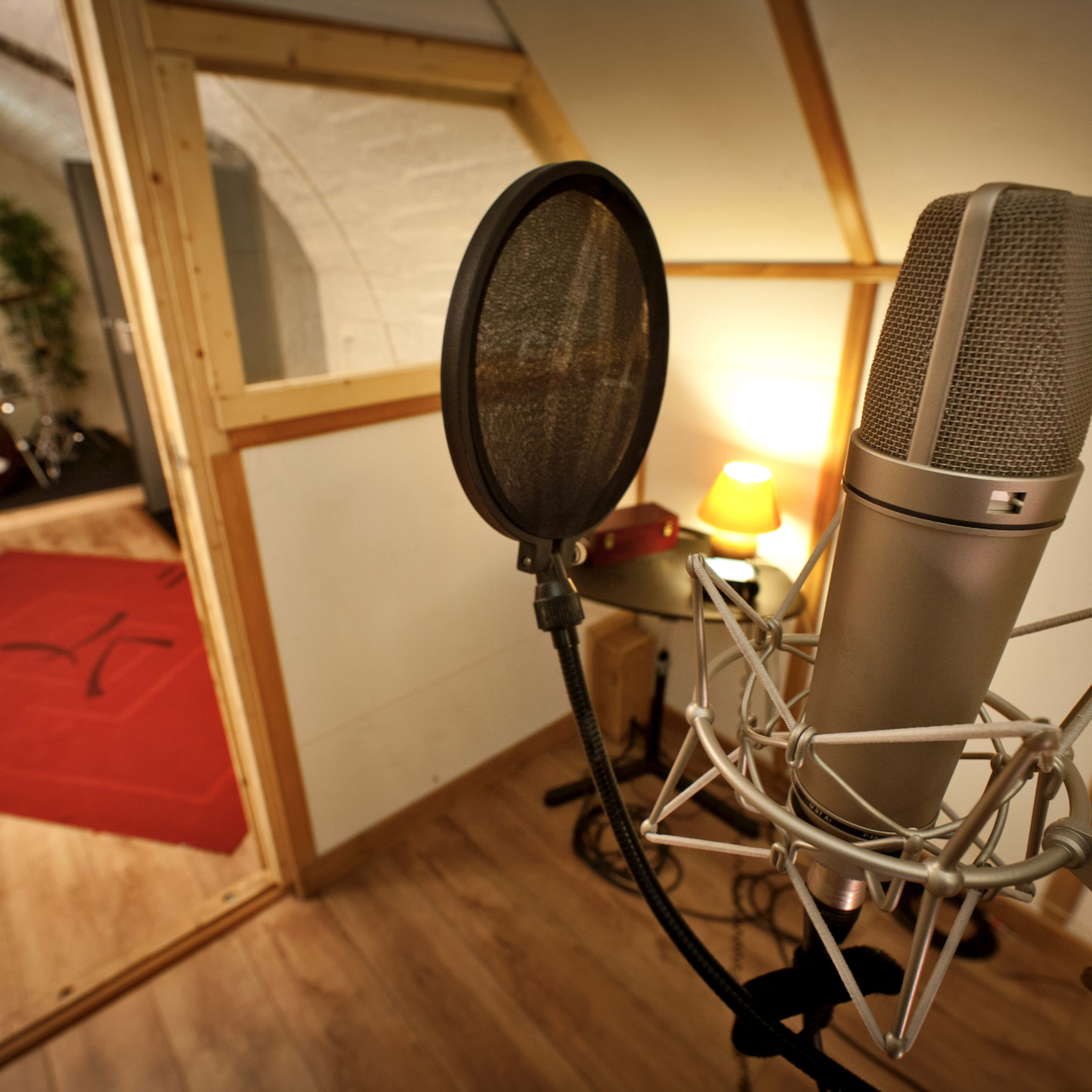 A microphone stands in the foreground with a soundproof booth and a drum set visible in the background.