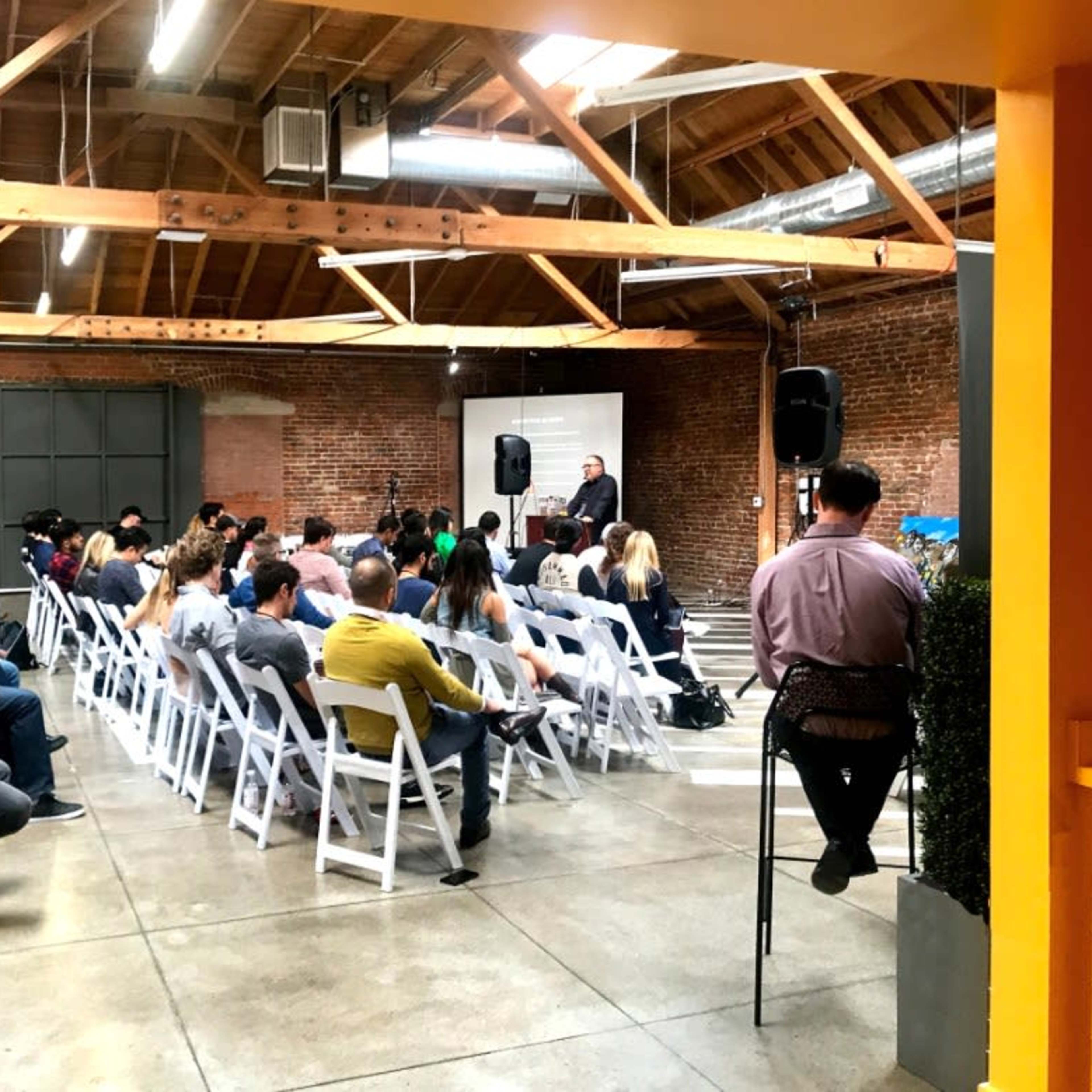 A speaker addresses an audience seated in rows at a well-lit event space with exposed brick walls and wooden beams.