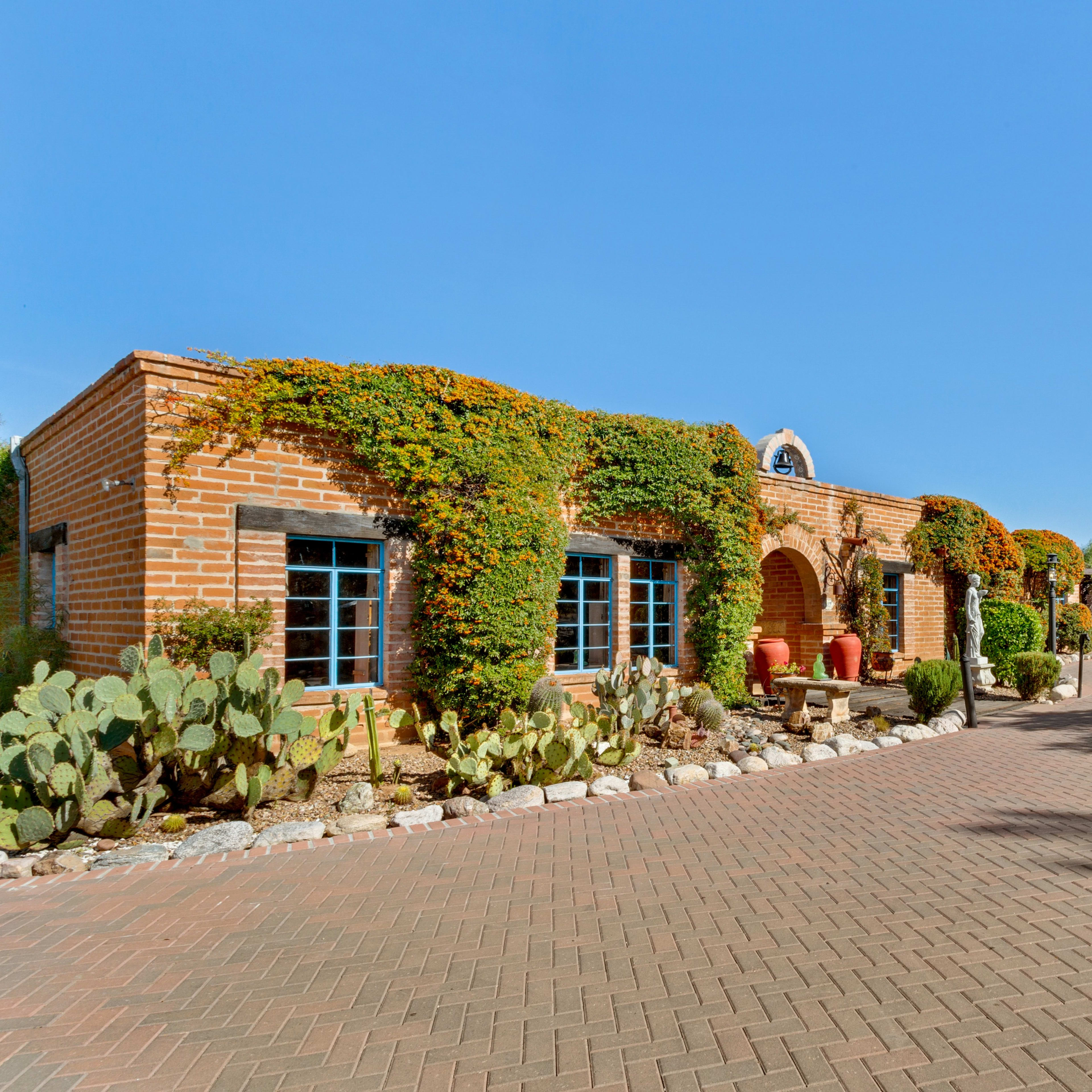 A brick house with green vines and blue windows is set along a paved driveway, leading to an adjoining building with a wooden door.