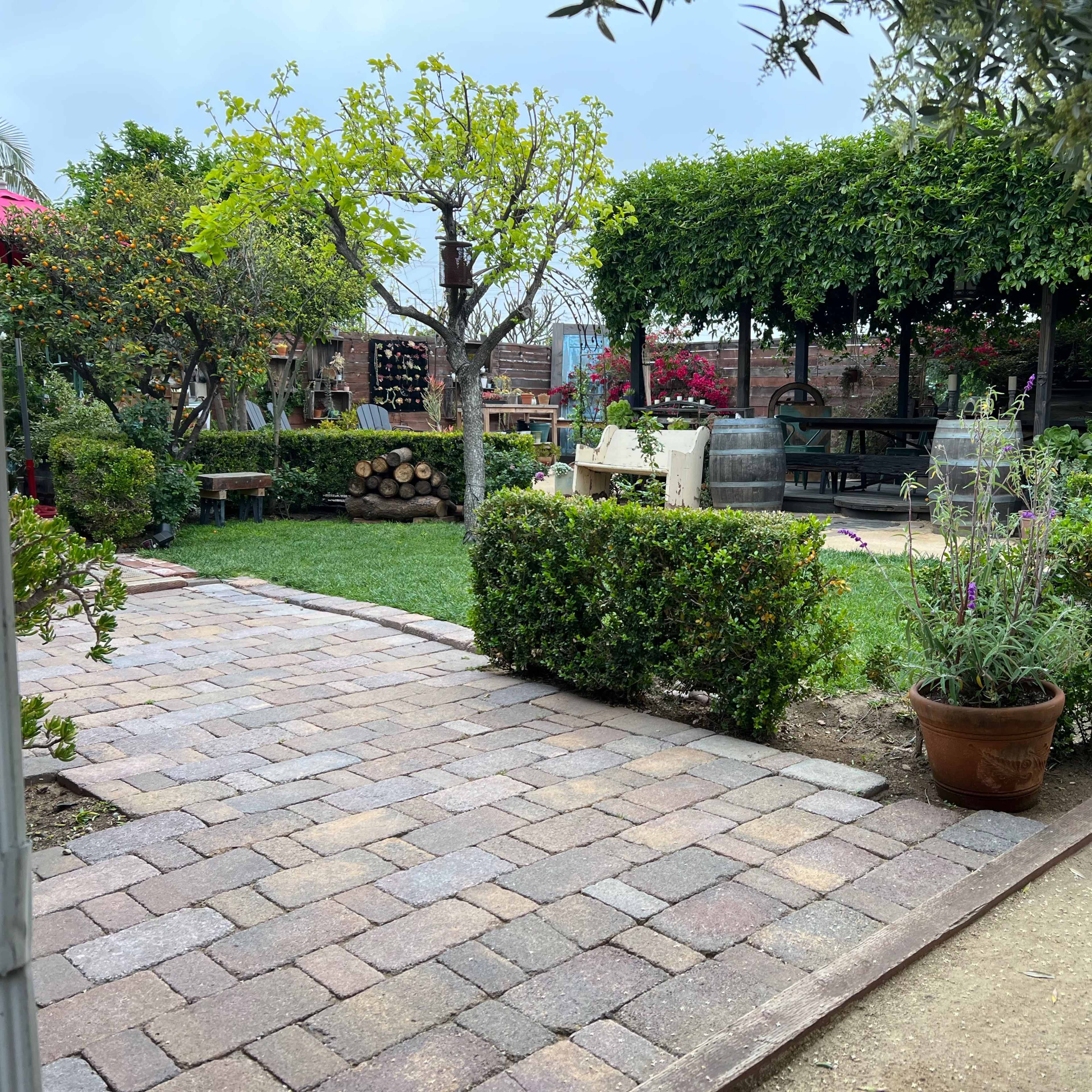 A brick pathway leads through a garden with lush greenery, potted plants, and shaded seating areas.
