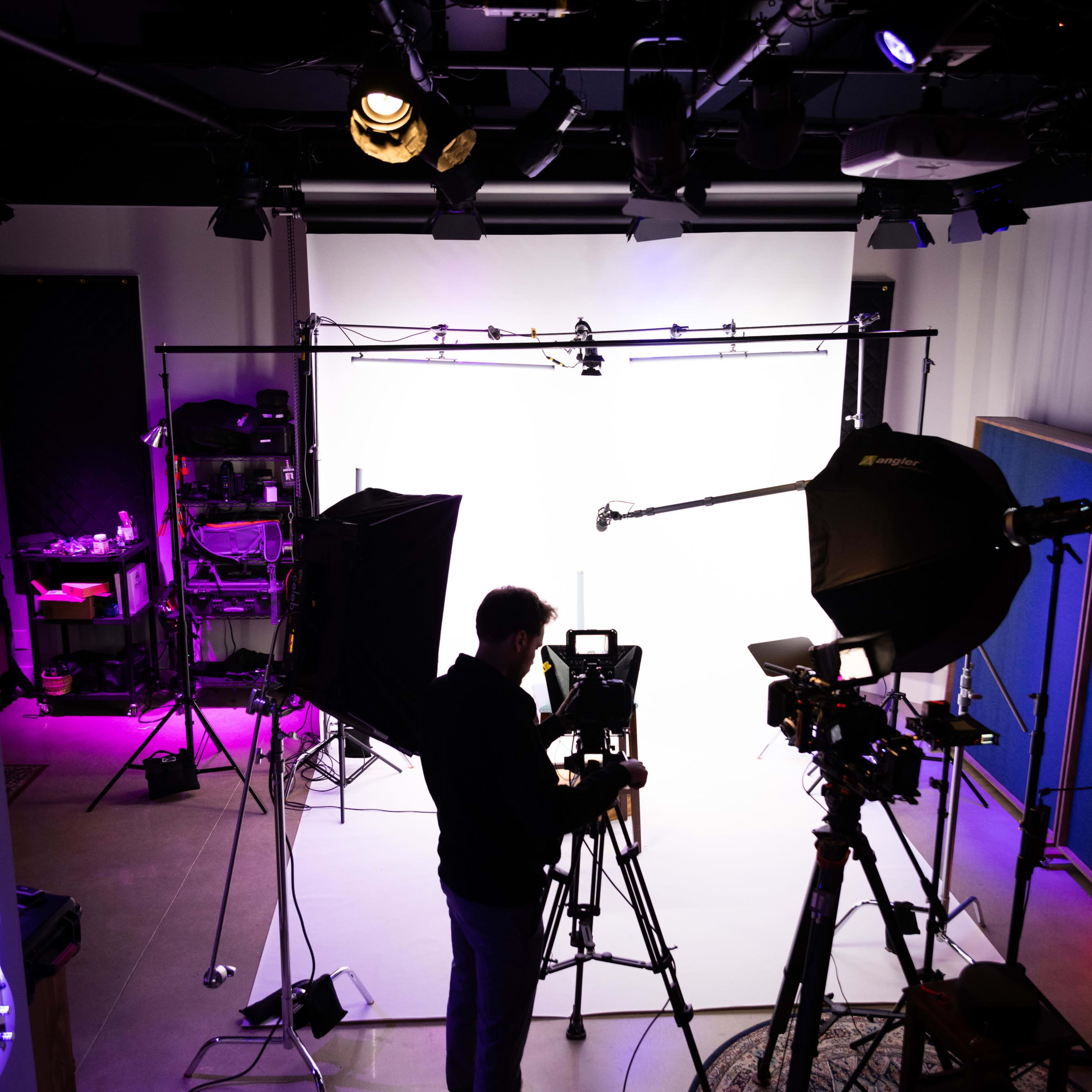 A person operates a camera in a well-equipped studio, surrounded by lighting setups and a white backdrop.