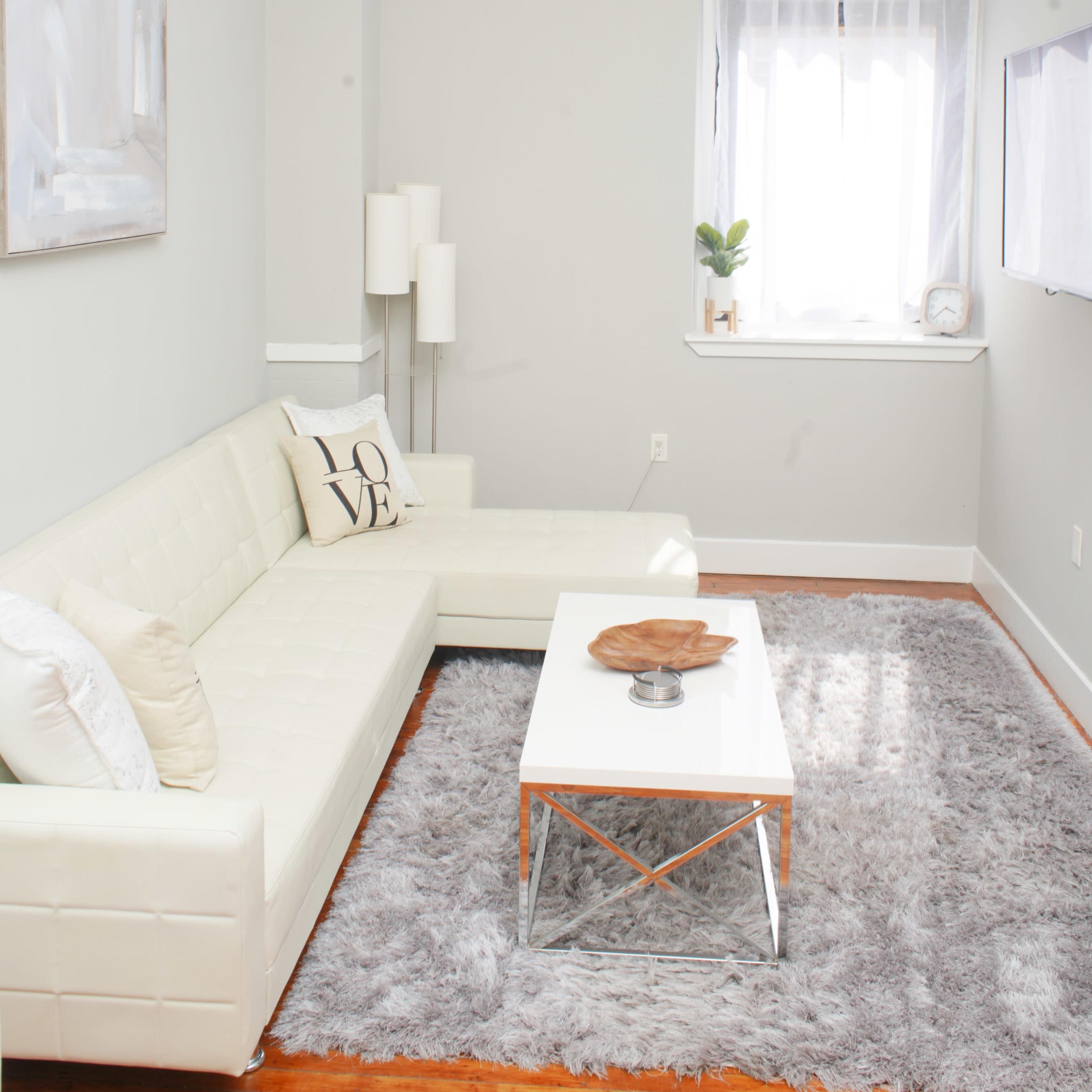 A modern living room features a white leather sectional sofa, a glass coffee table, and a large area rug, with a window allowing natural light to enter.