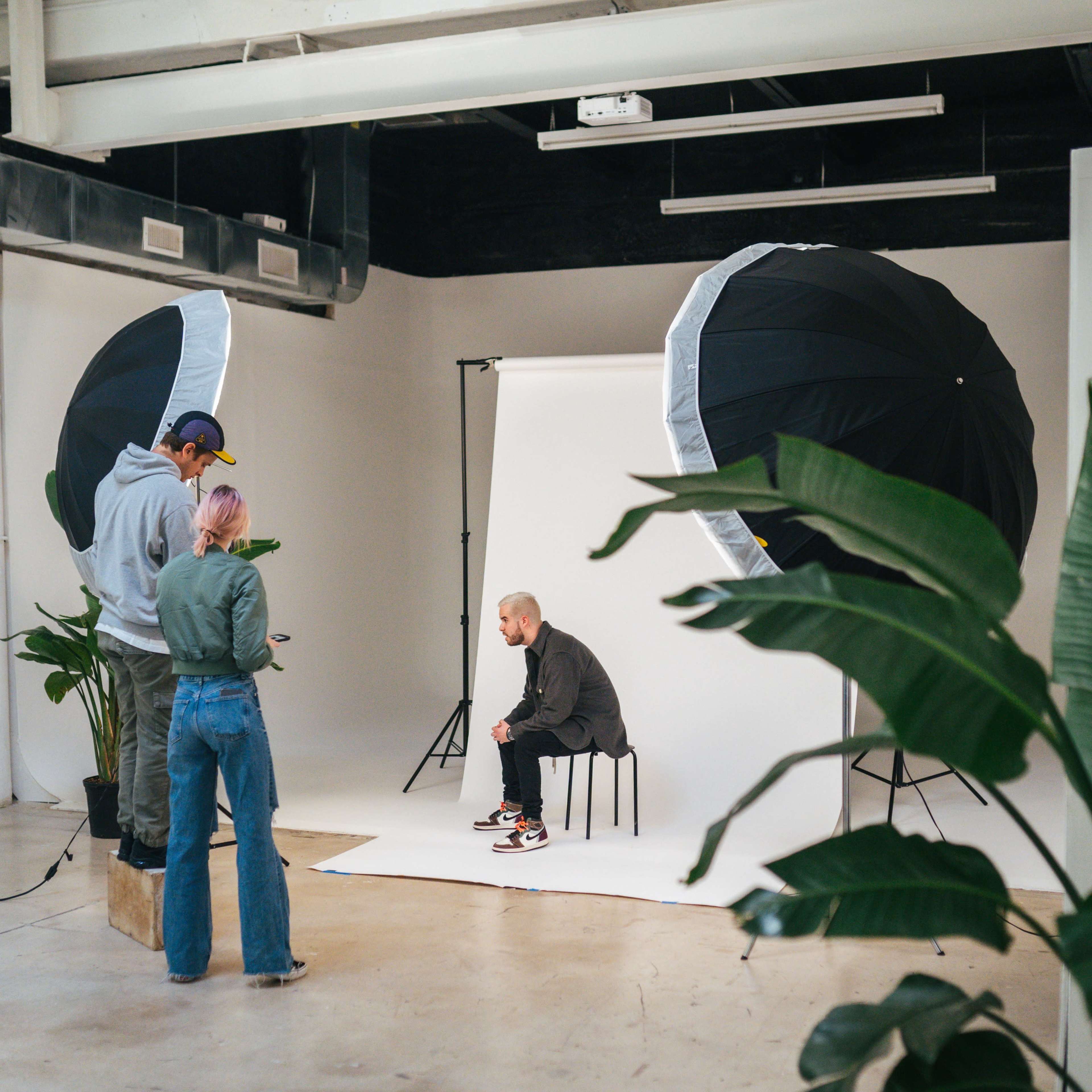 A male model sits on a stool in a photo studio, while a stylist adjusts the scene and a photographer prepares to take a picture.