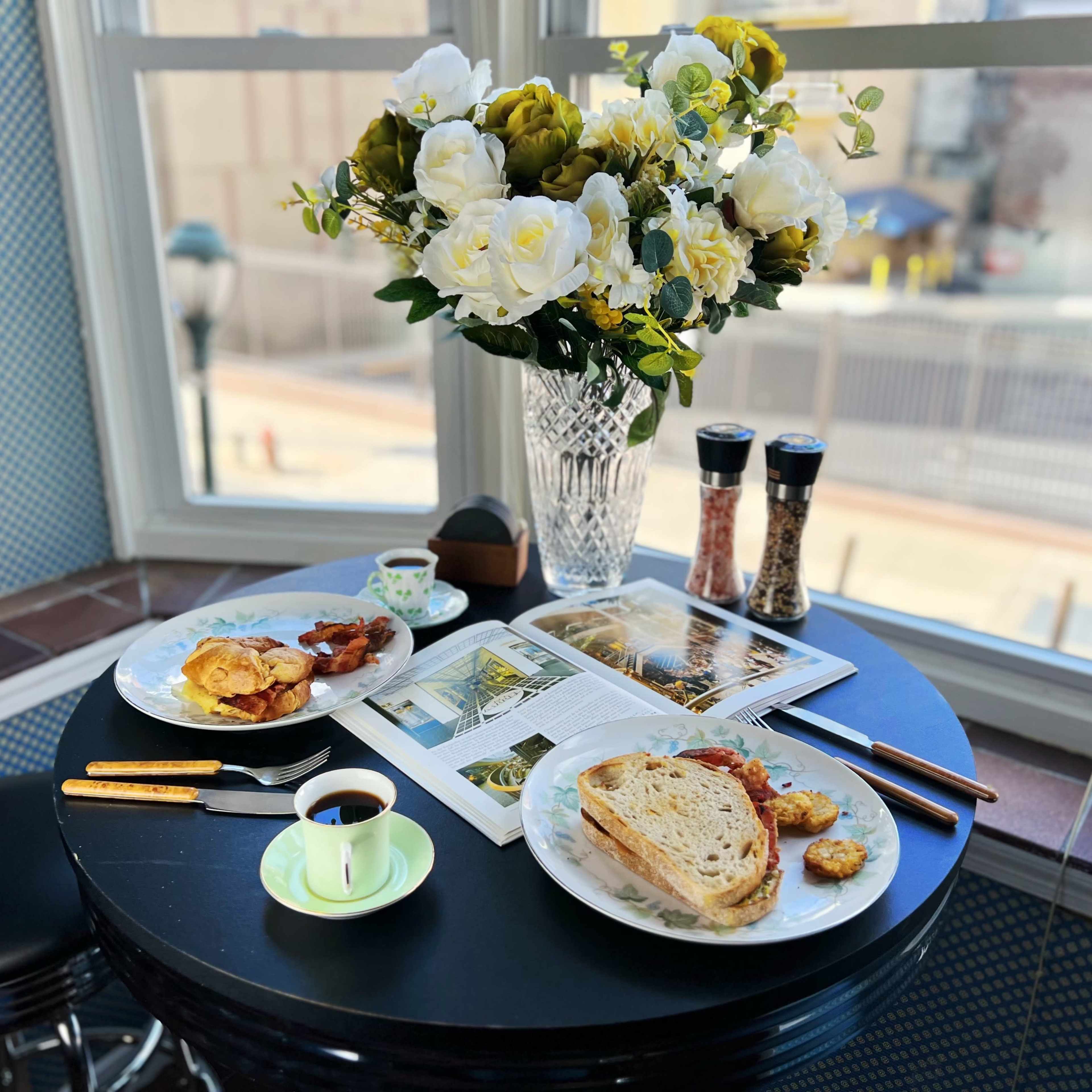 A breakfast setting features plates of food, a cup of coffee, and a vase of flowers on a round table near a window.