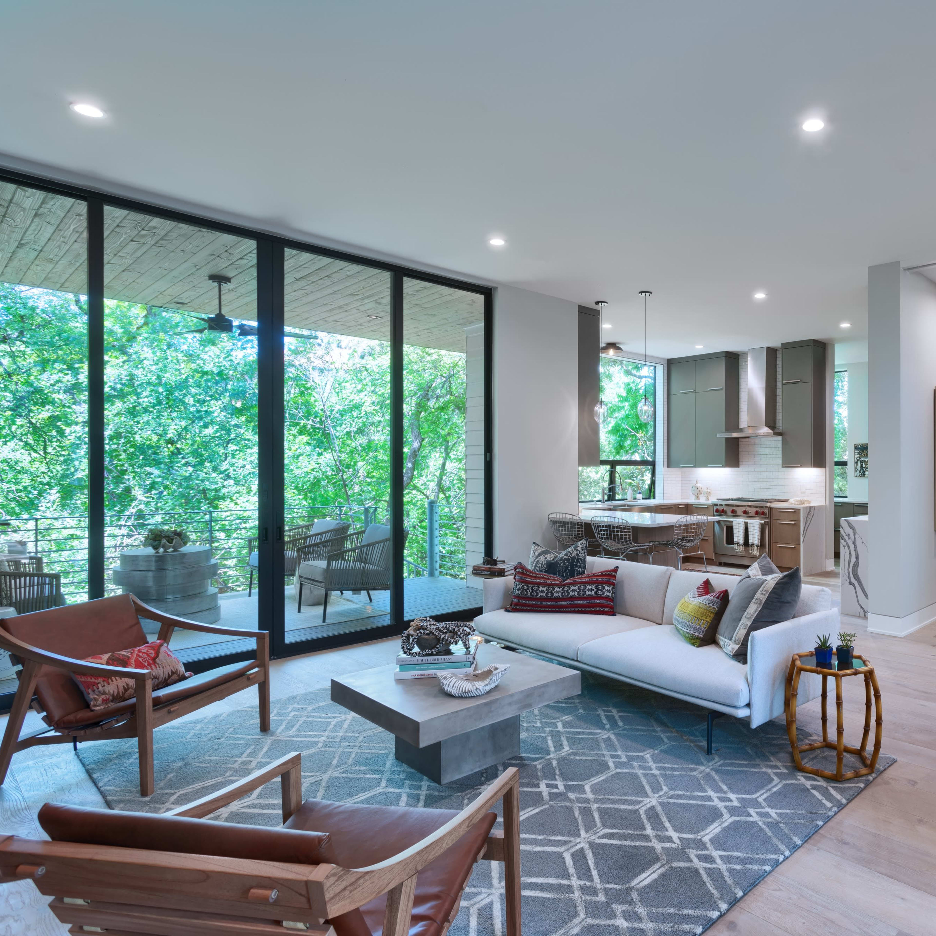 A modern living room with large windows overlooking greenery, featuring a seating area, a dining space, and a kitchen in the background.
