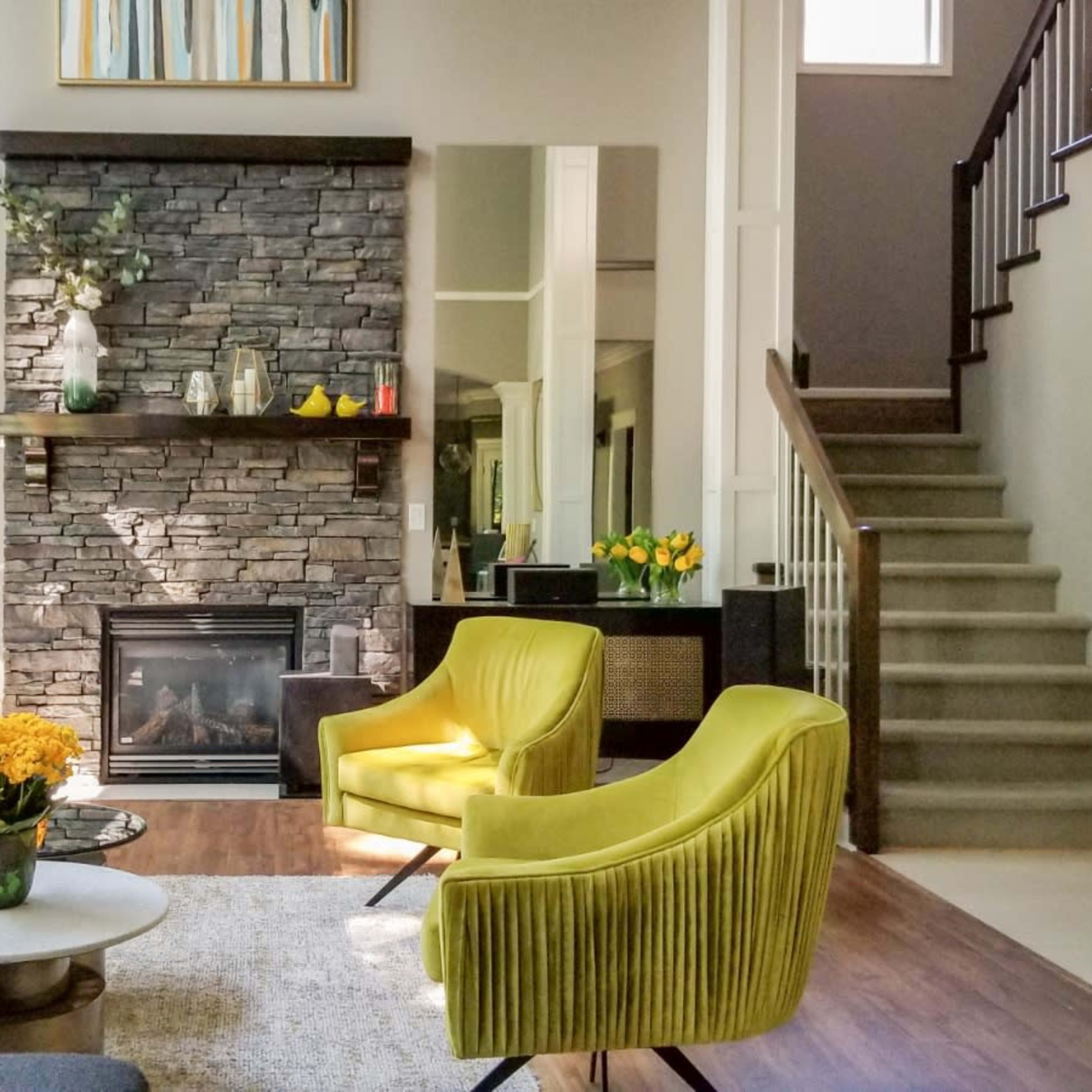 A modern living room with large windows, a stone fireplace, and yellow accent chairs, adjacent to a staircase.