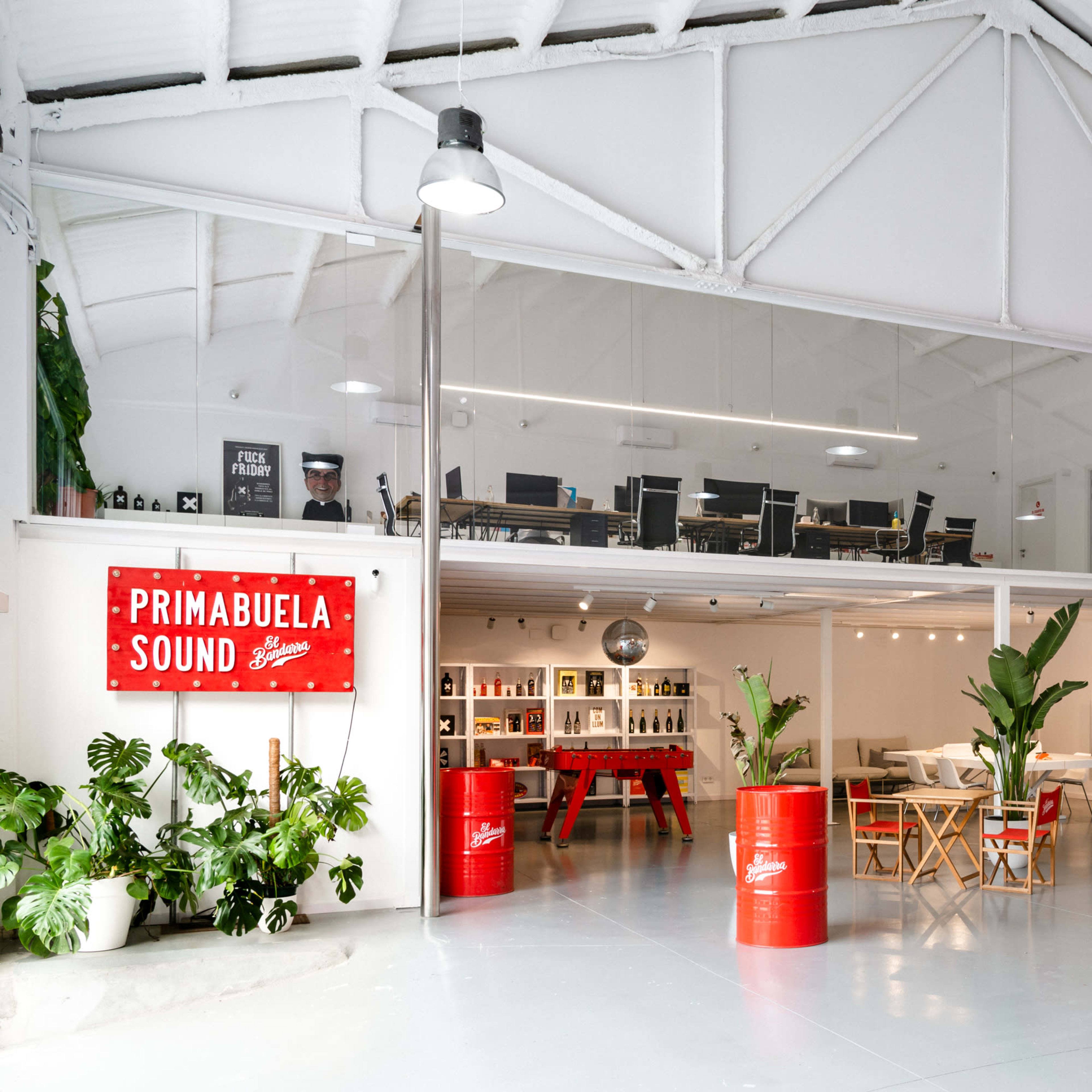 A modern office space with an upstairs area, featuring red barrels as planters, a sign reading "Primabuela Sound," and people working in an open layout.