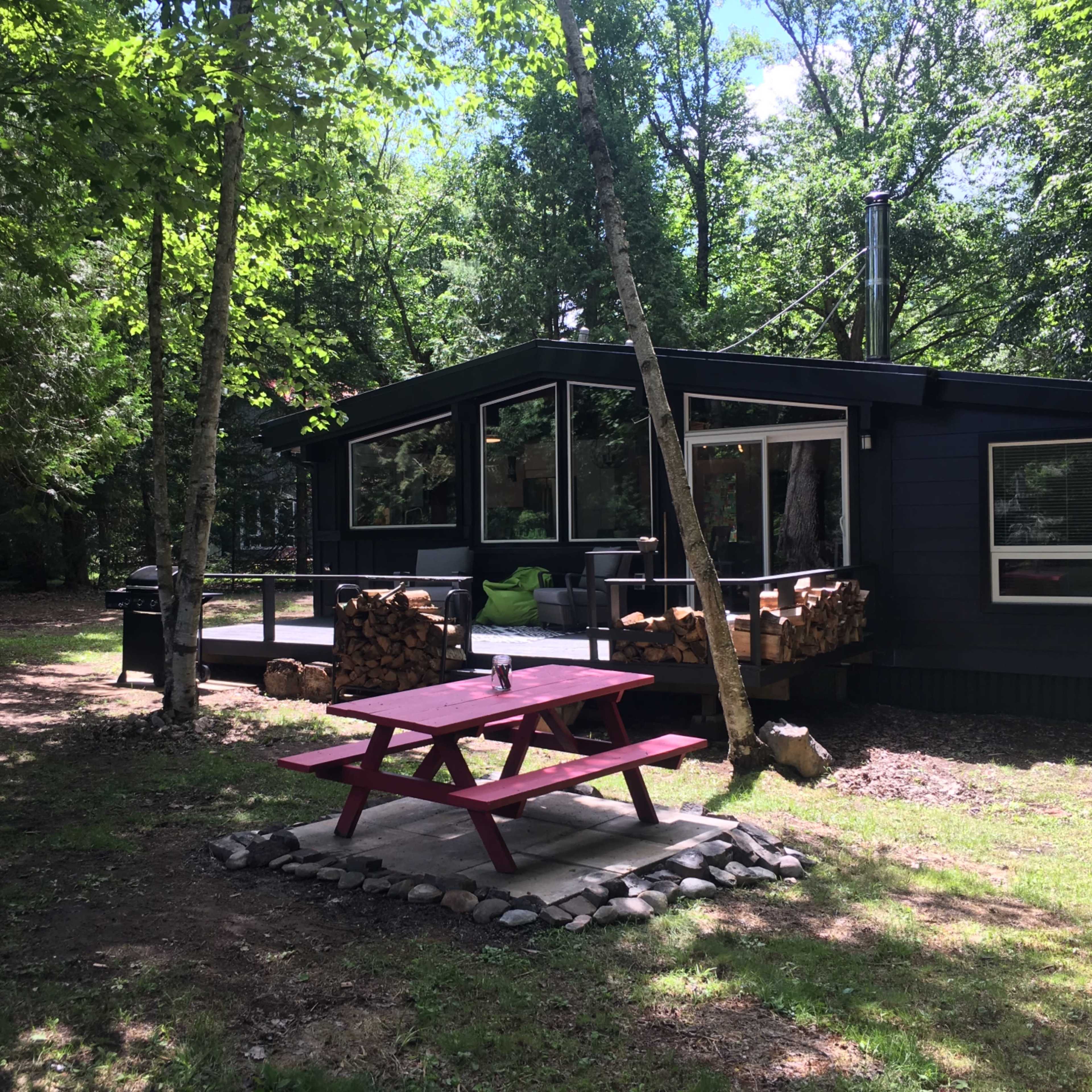 A red picnic table sits on a stone circle in front of a modern black cabin surrounded by trees.