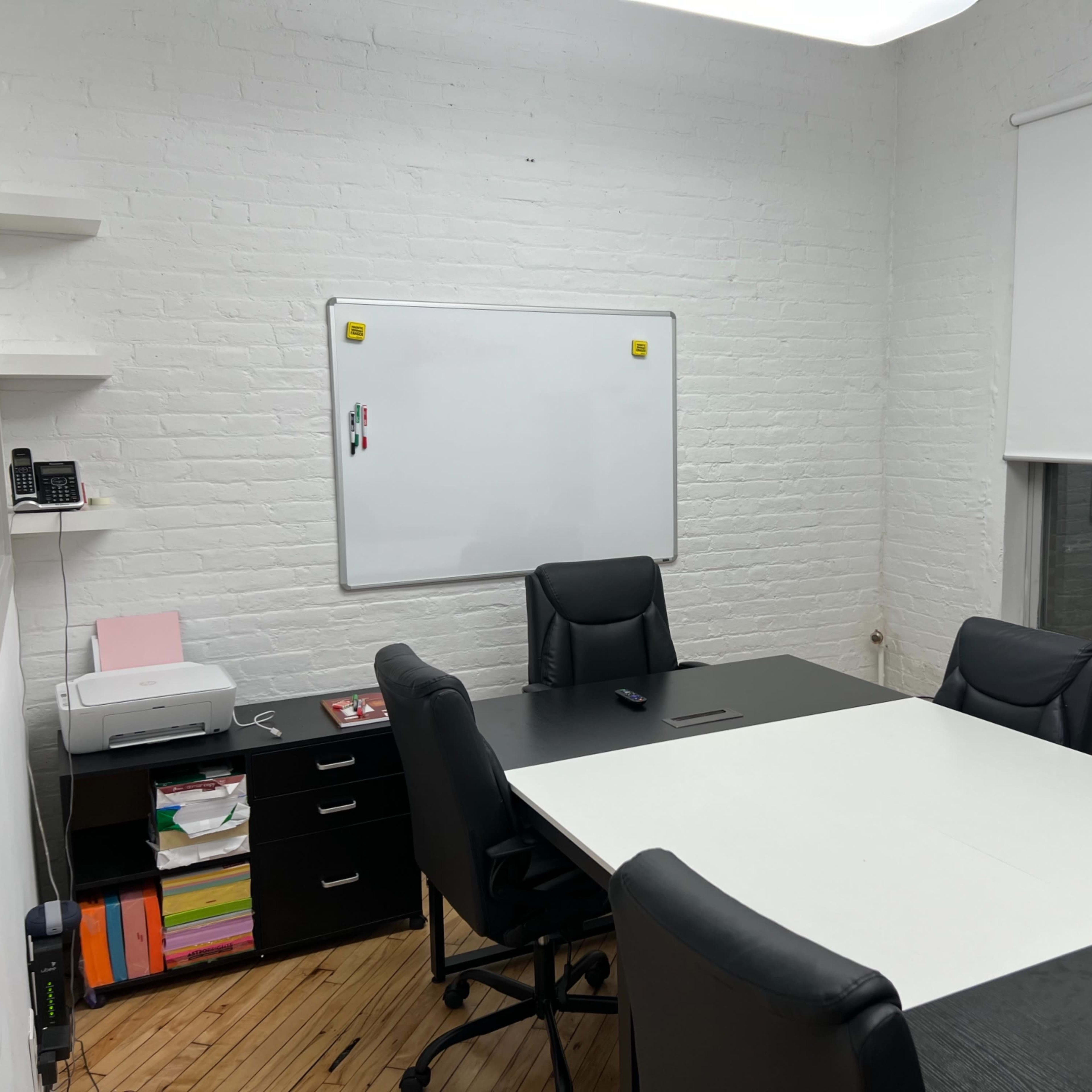 A simple conference room featuring a black table, black chairs, a whiteboard on the wall, and a printer on a nearby shelf.