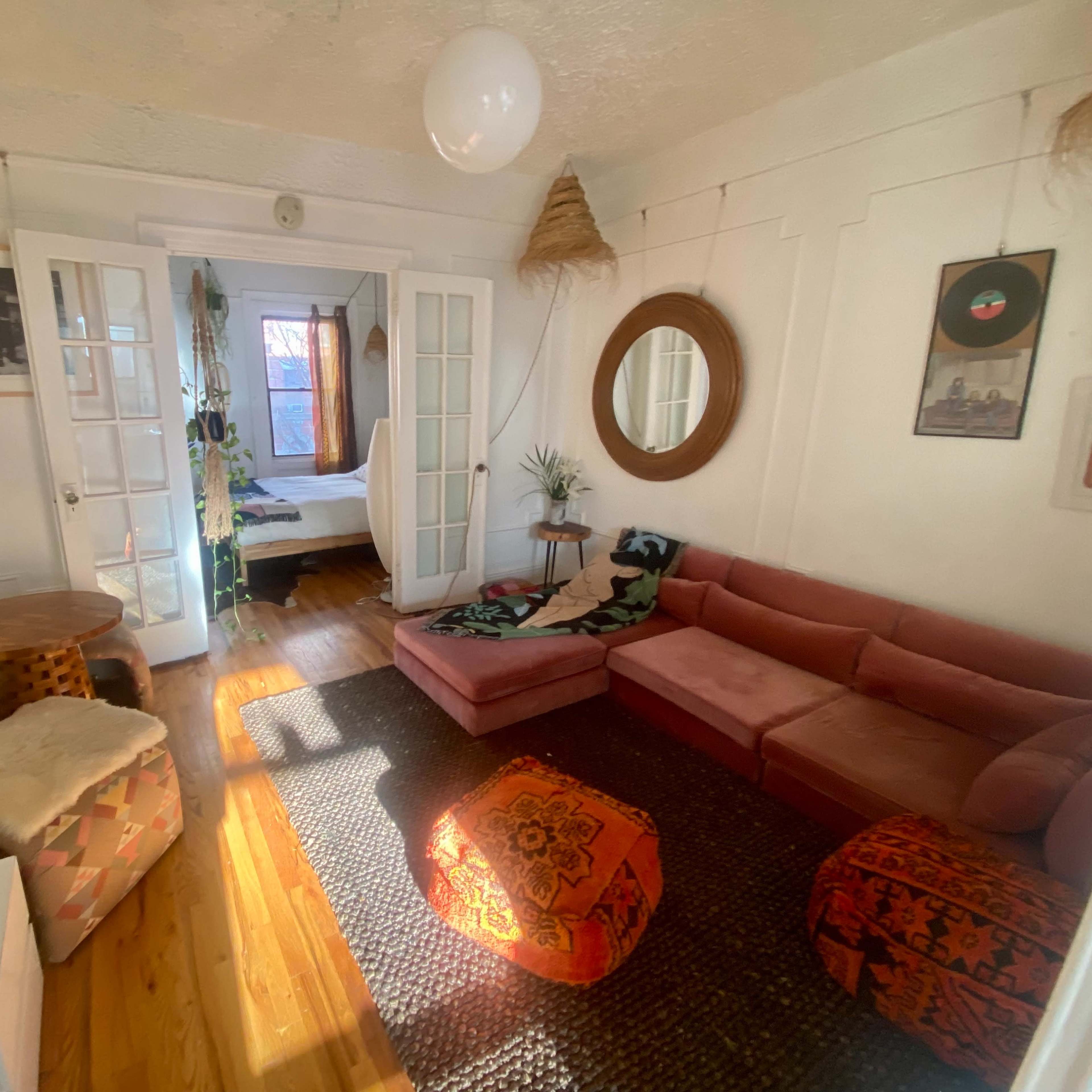 A cozy living room featuring a pink sofa, decorative poufs, a round mirror, and wooden flooring, with a view into a bedroom through glass doors.