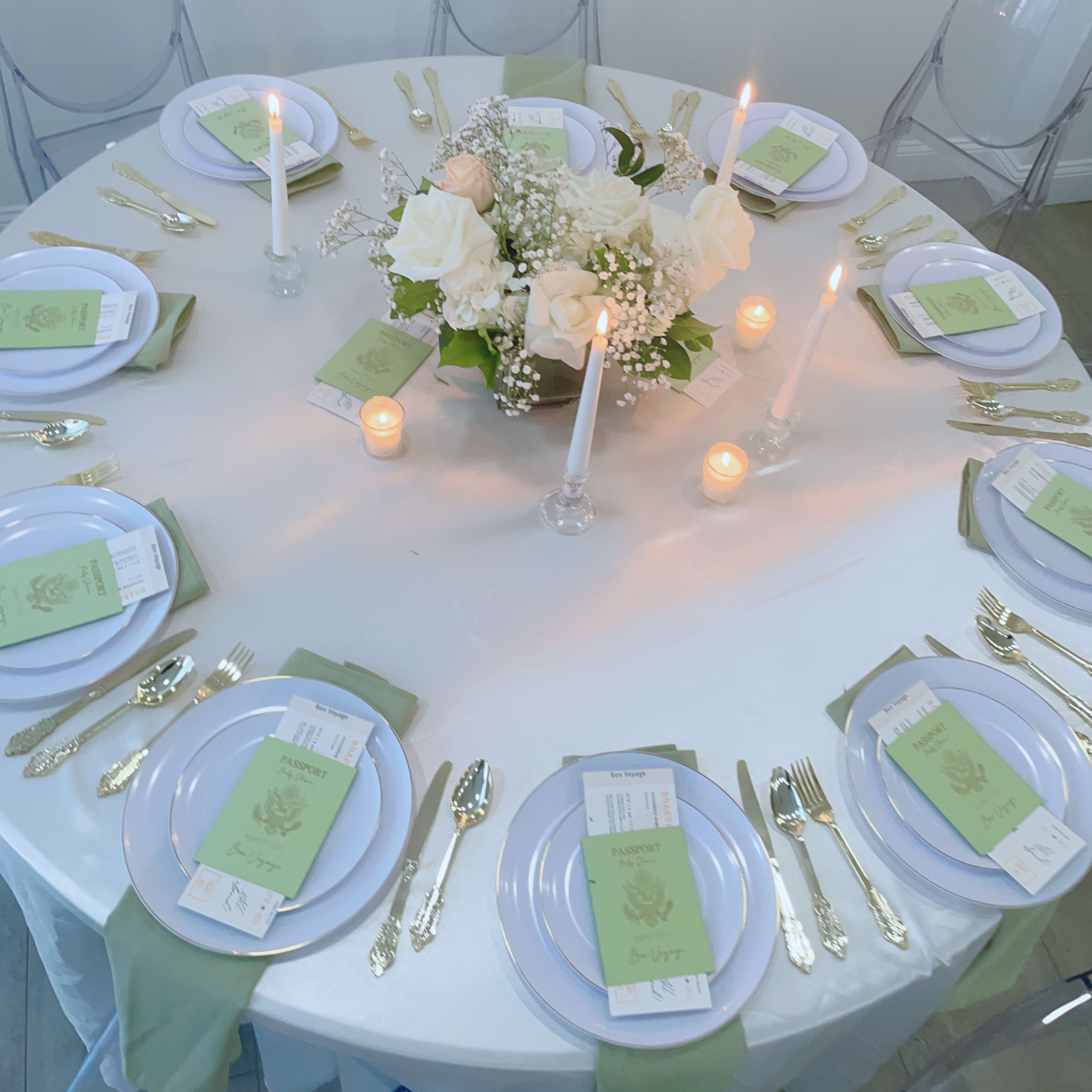 A round dining table is set with white plates, green menus, gold cutlery, and a centerpiece of white roses and candles.