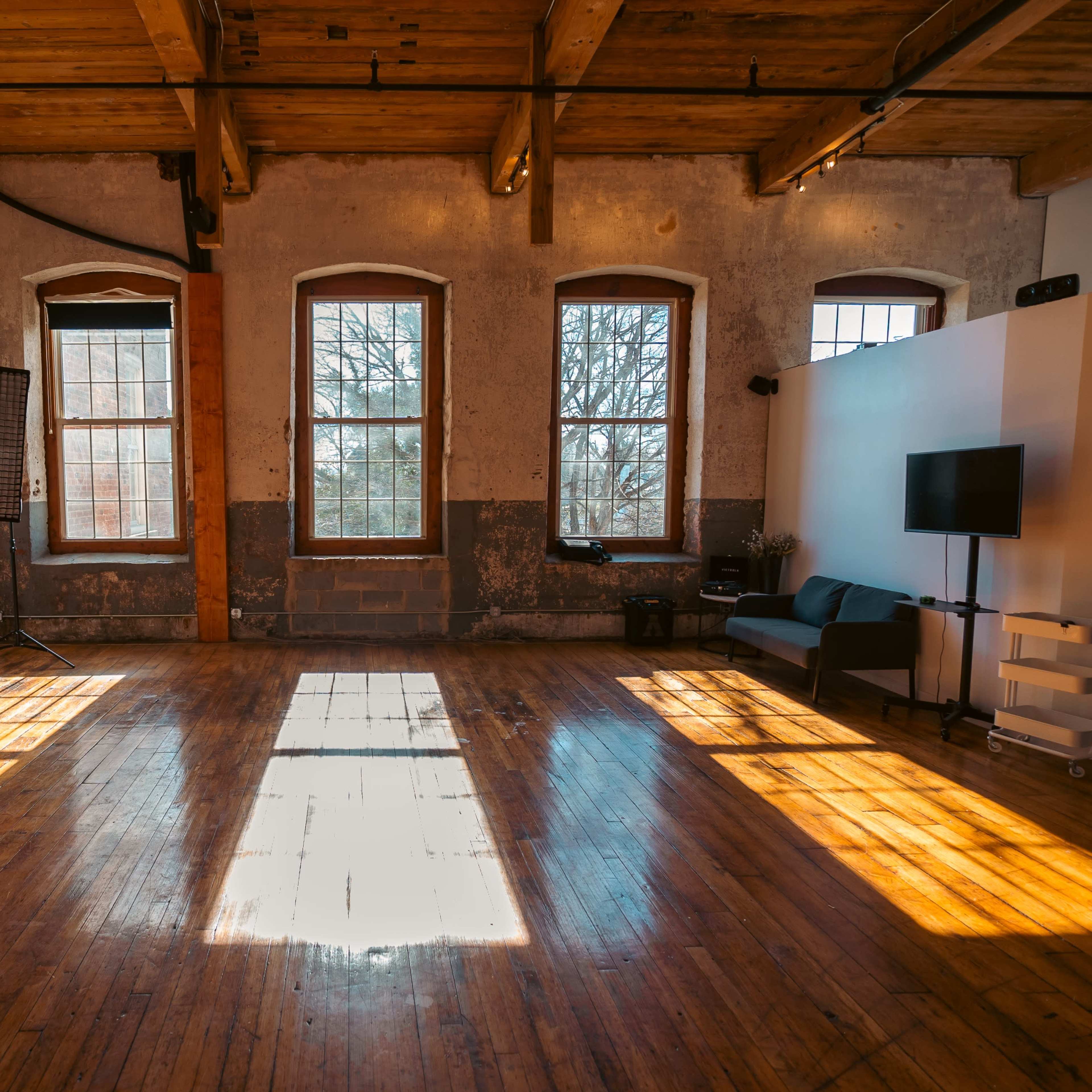 A spacious, well-lit studio with wooden floors, large windows, and a seating area.