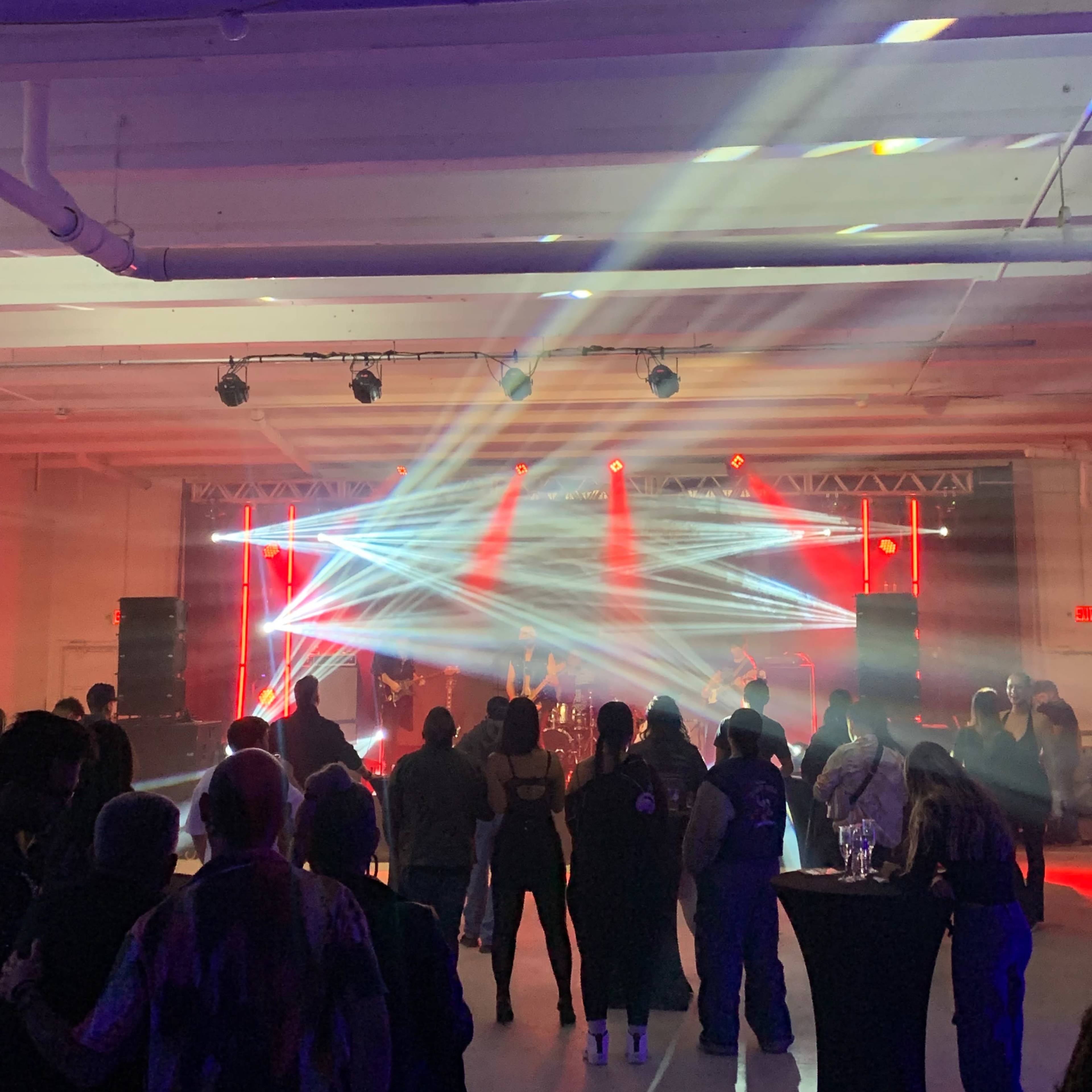 A crowd watches a band perform on stage with colorful lights and fog effect in a spacious indoor venue.