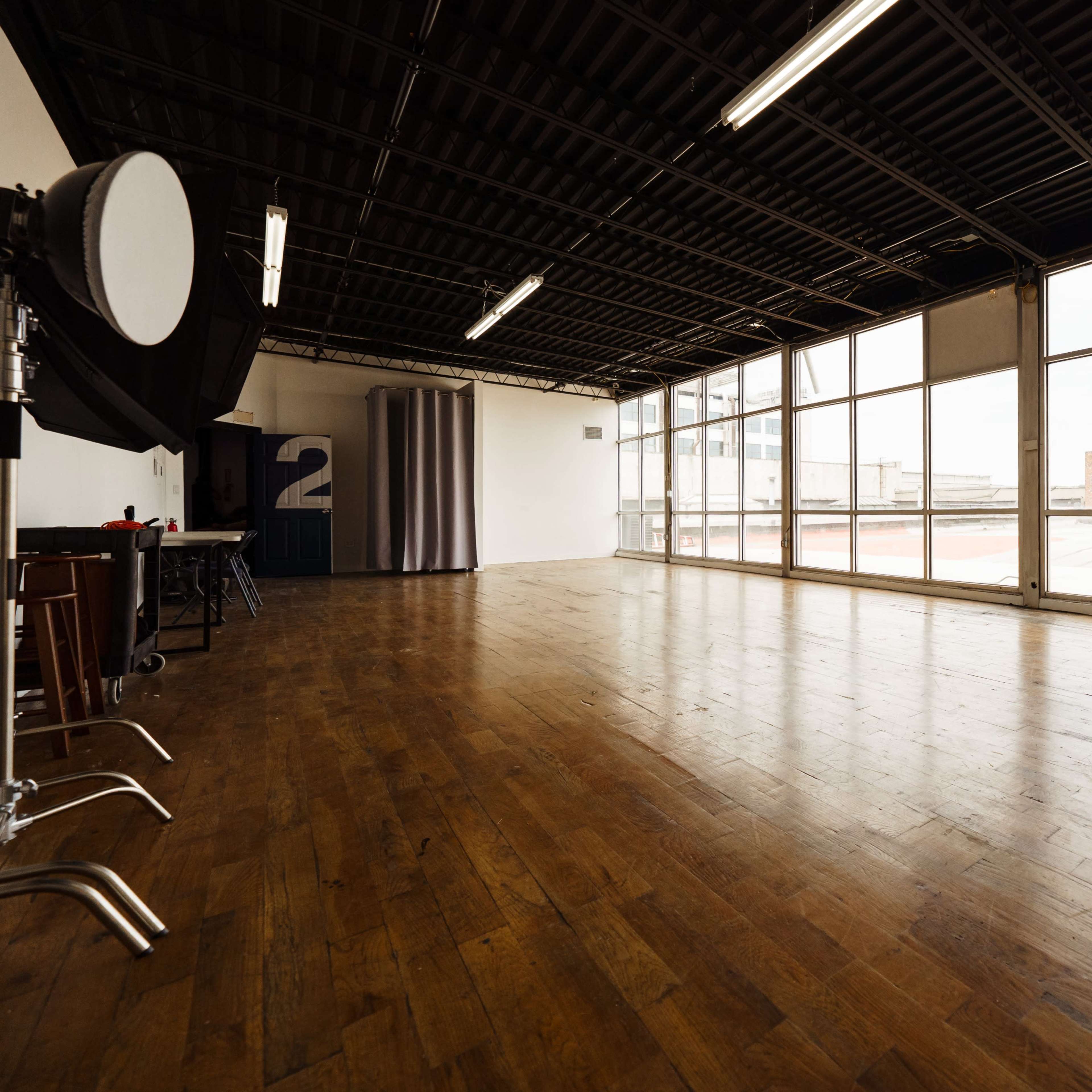 A spacious, empty studio with wooden flooring and large windows allowing natural light to enter.