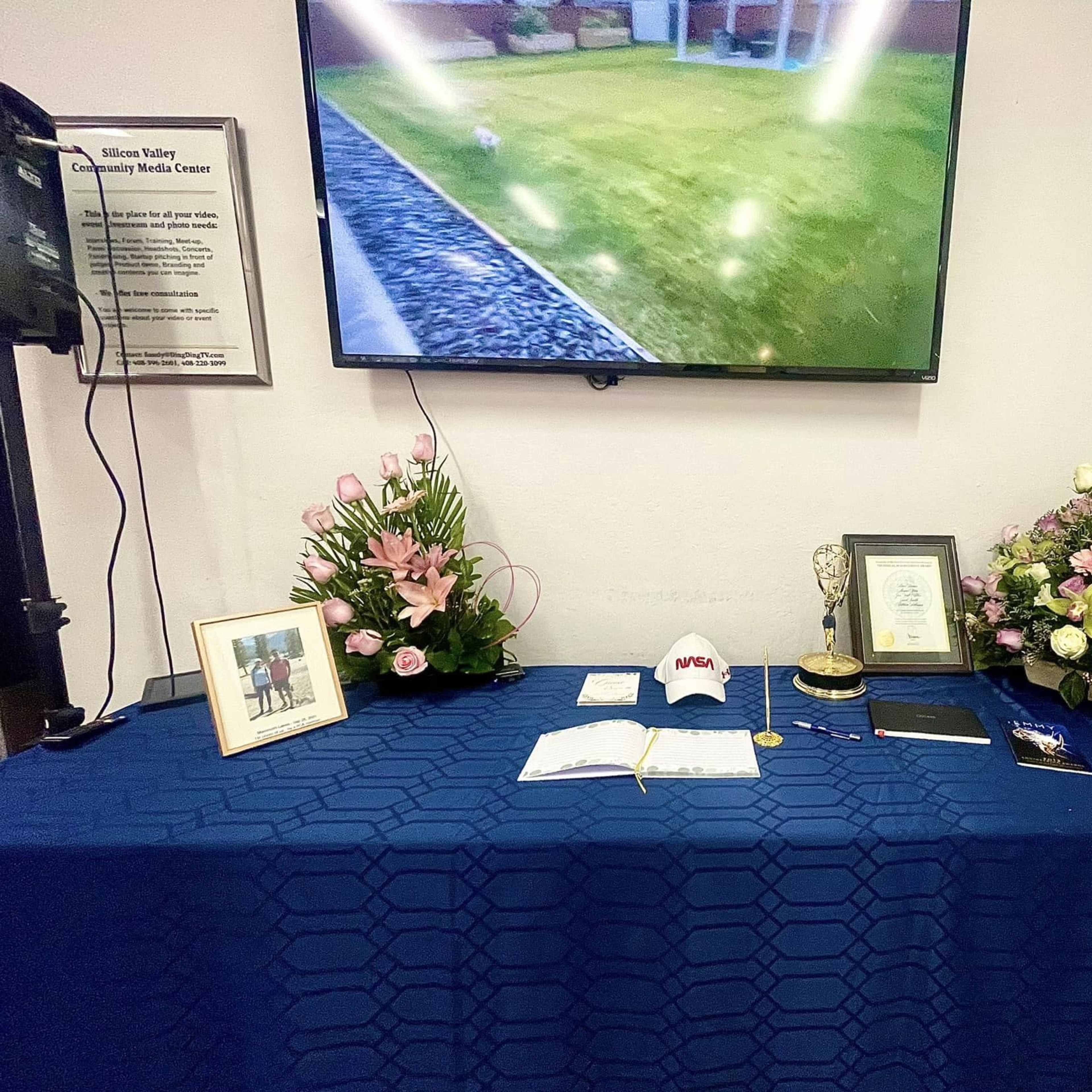 A display table featuring a blue tablecloth, framed photos, a trophy, flowers, a notebook, and a cap, with a TV mounted on the wall showing a garden view.