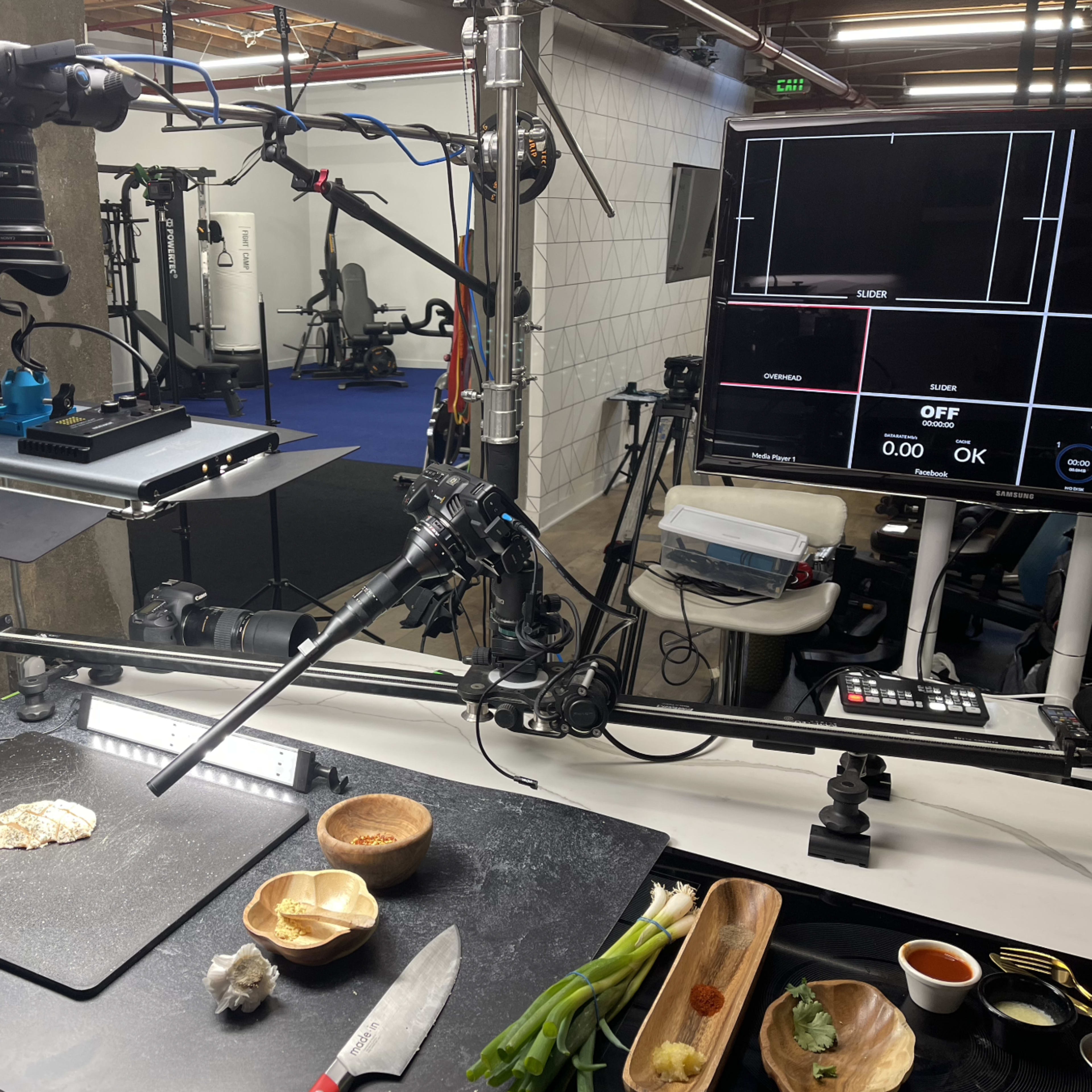 A food photography setup with a camera mounted on a multi-arm rig, capturing ingredients and utensils arranged on a dark countertop.