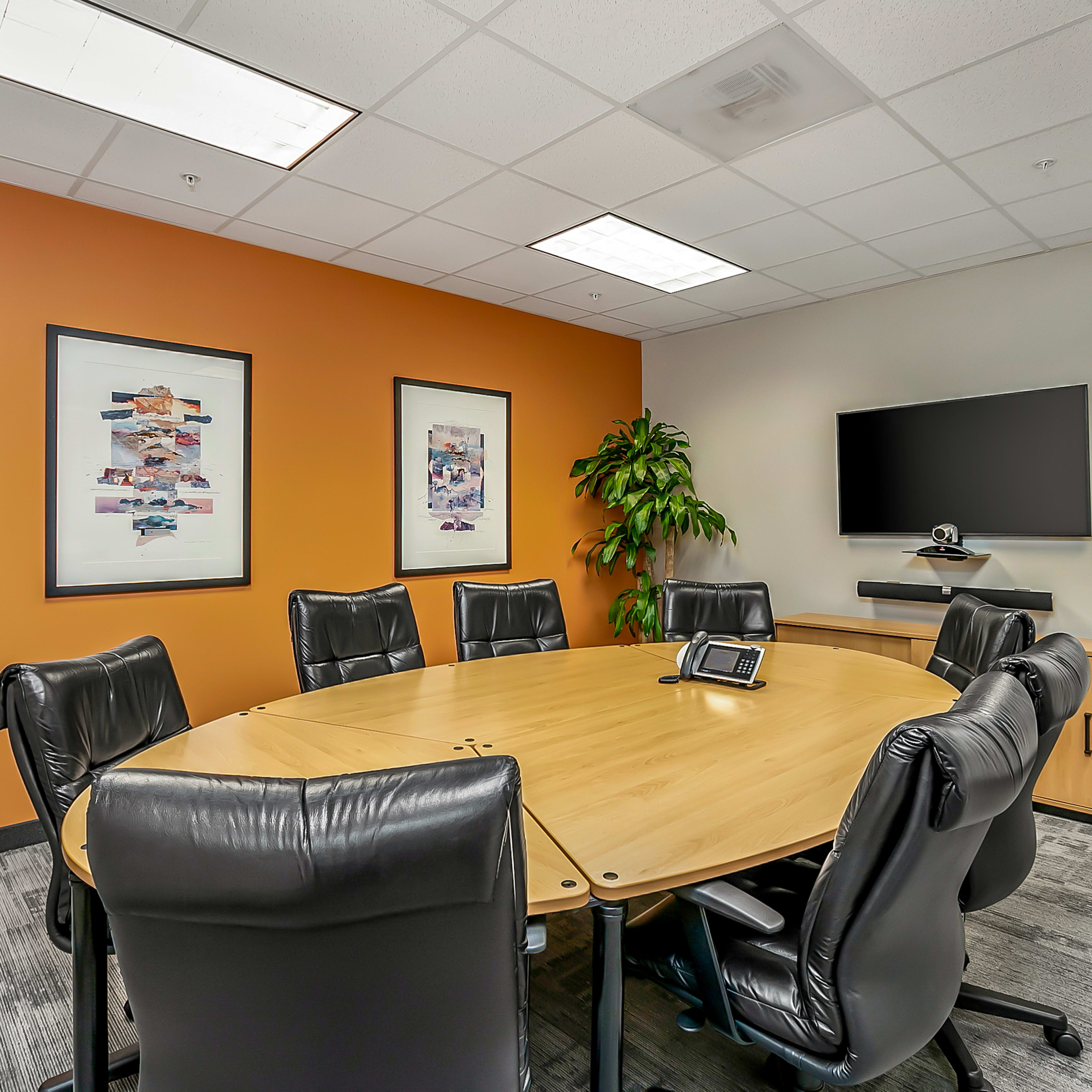 A conference room features a large round wooden table surrounded by black leather chairs, with art on the walls, plants in the corners, and a wall-mounted TV.