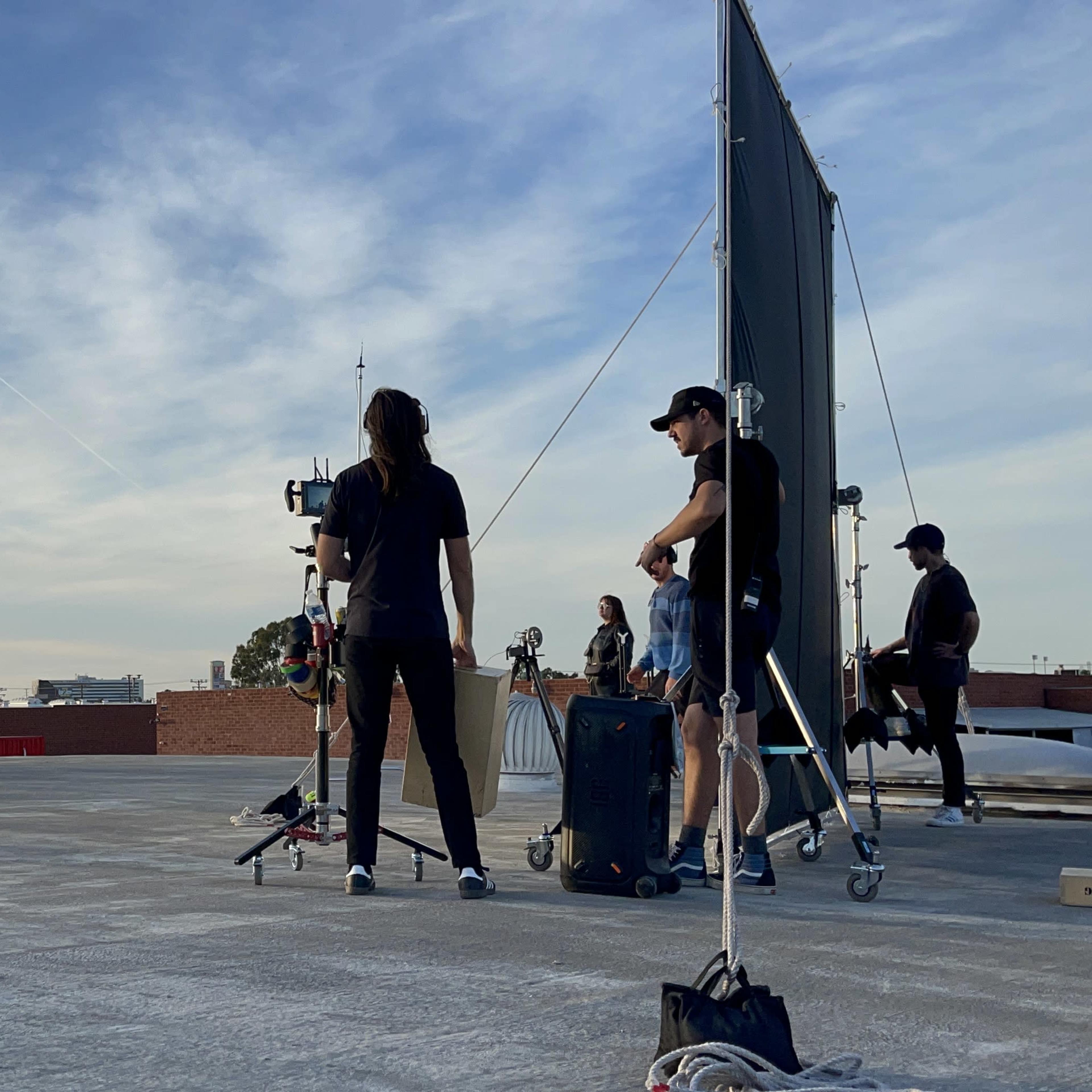 A group of people are gathered on a rooftop, while a crew sets up equipment and adjusts a backdrop for a photoshoot.