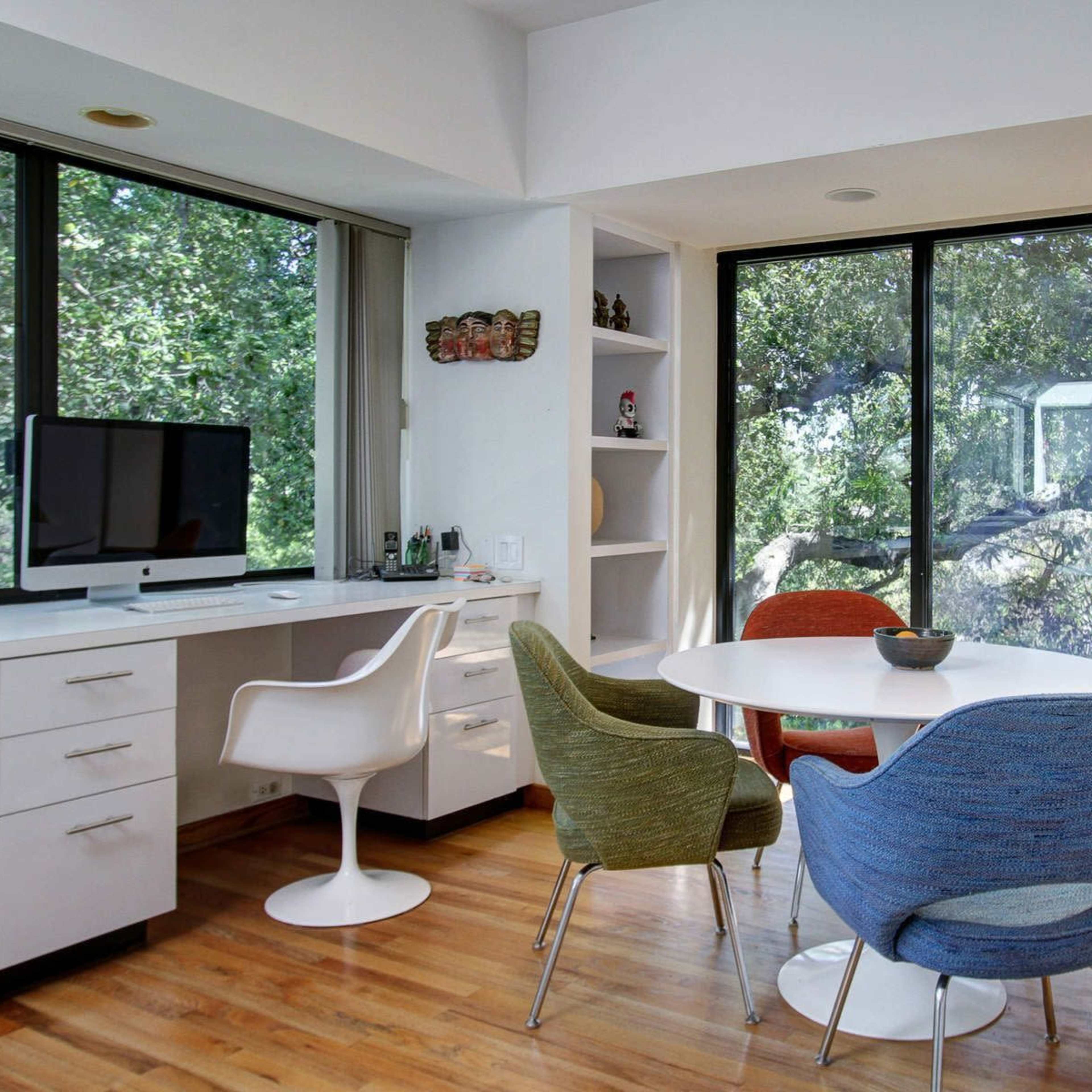 A modern dining area features a round table surrounded by colorful chairs, with a desktop computer and large windows overlooking greenery.